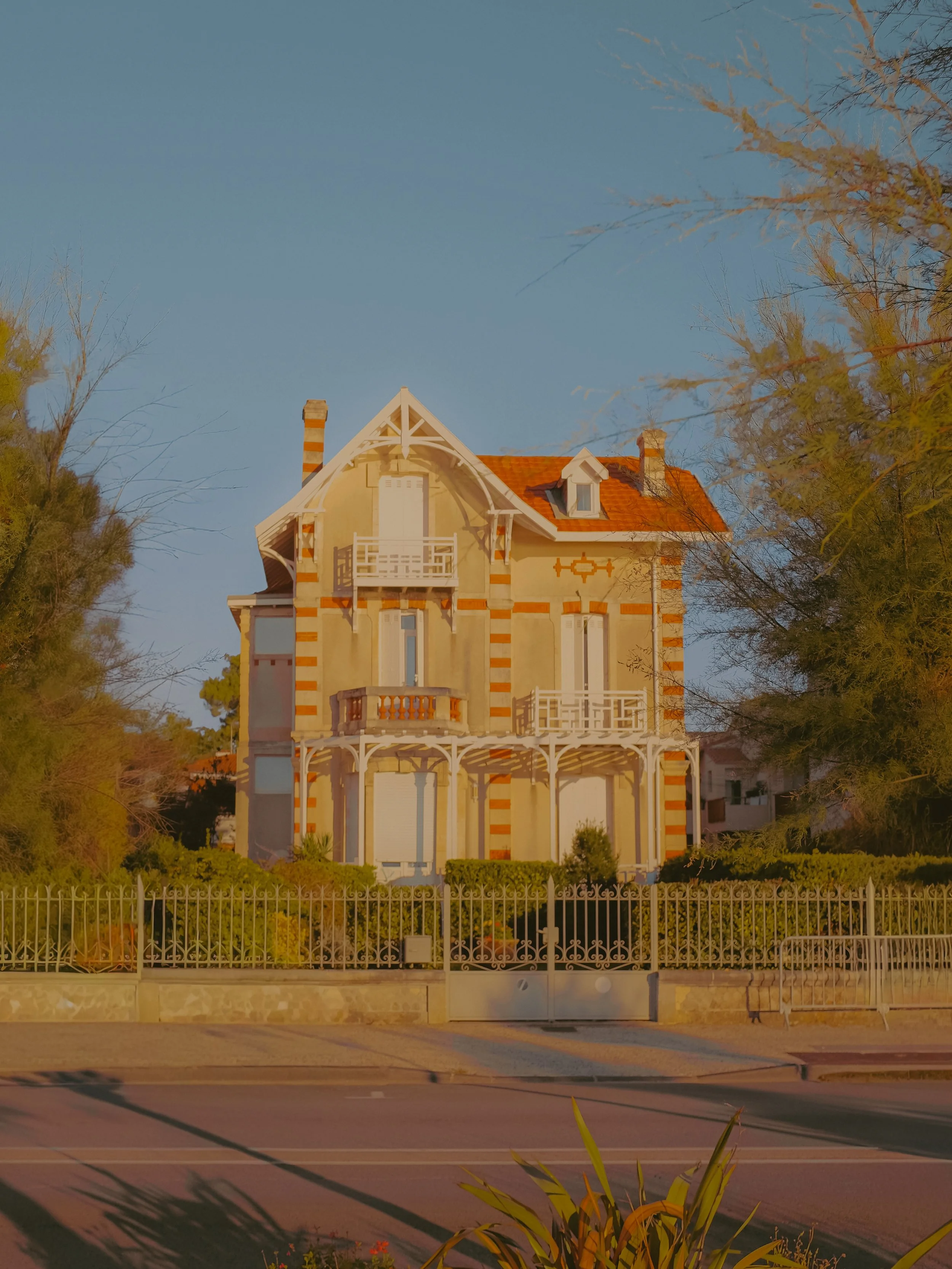 Une maison elegante avec une façade jaune, des détails en blanc, un balcon, entourée de verdure, sous un ciel bleu.
