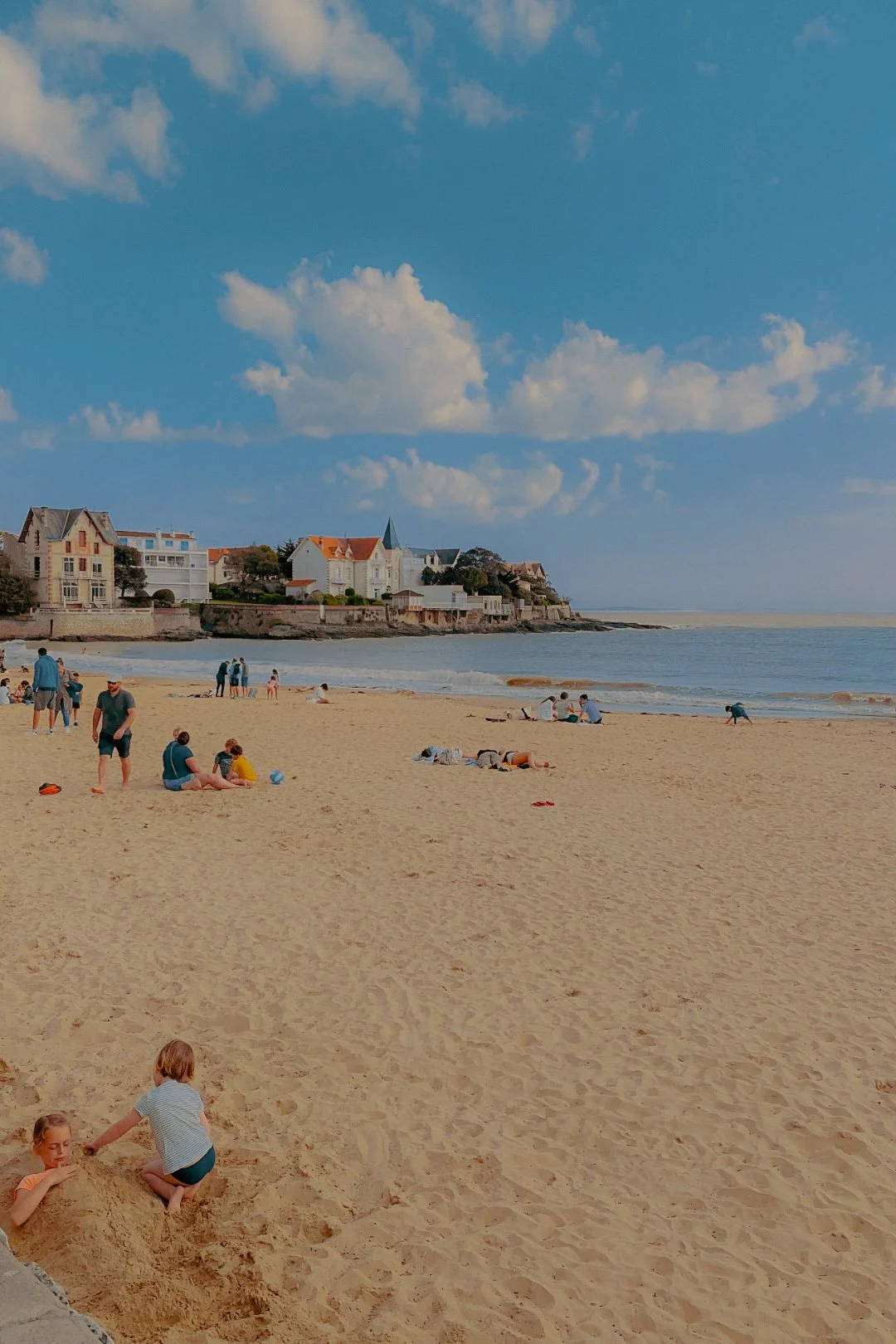 Plage avec des gens se relaxant, jouant dans le sable, et se promenant près de la mer, avec un village de maisons en arrière-plan et un ciel bleu avec des nuages.