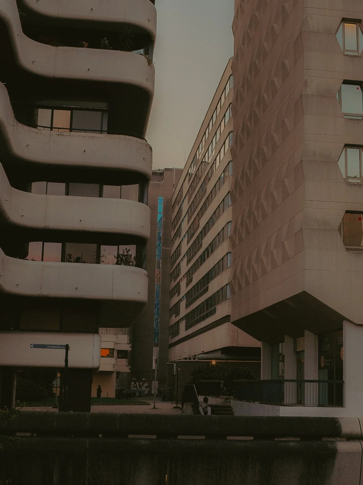 Vue urbaine avec deux immeubles modernes, un à gauche avec des balcons courbes et un à droite avec une façade en motifs géométriques, au crépuscule.