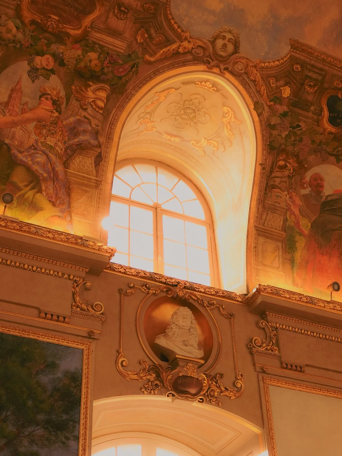 Intérieur rococo avec un grand plafond peint, une fenêtre lumineuse, un portrait de Louis XIV, et des décorations florales et dorées.