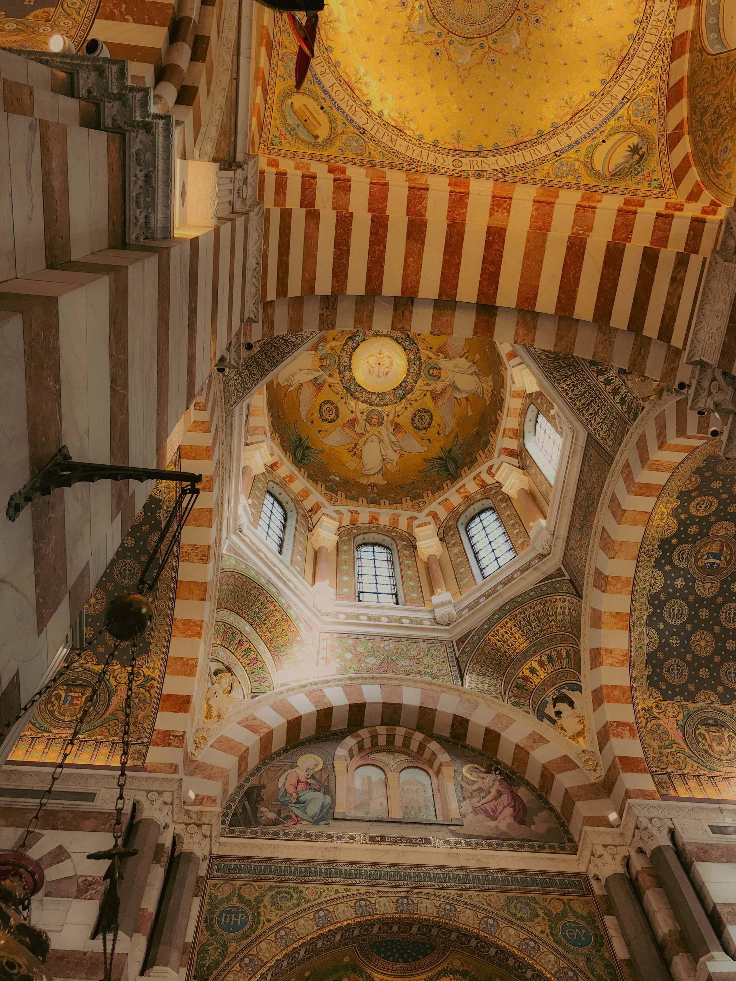Intérieur d'une église ornée de décorations colorées, avec des colonnes rayées rouge et blanc, des fenêtres en verre coloré, et un plafond orné de fresques et mosaïques dorées.