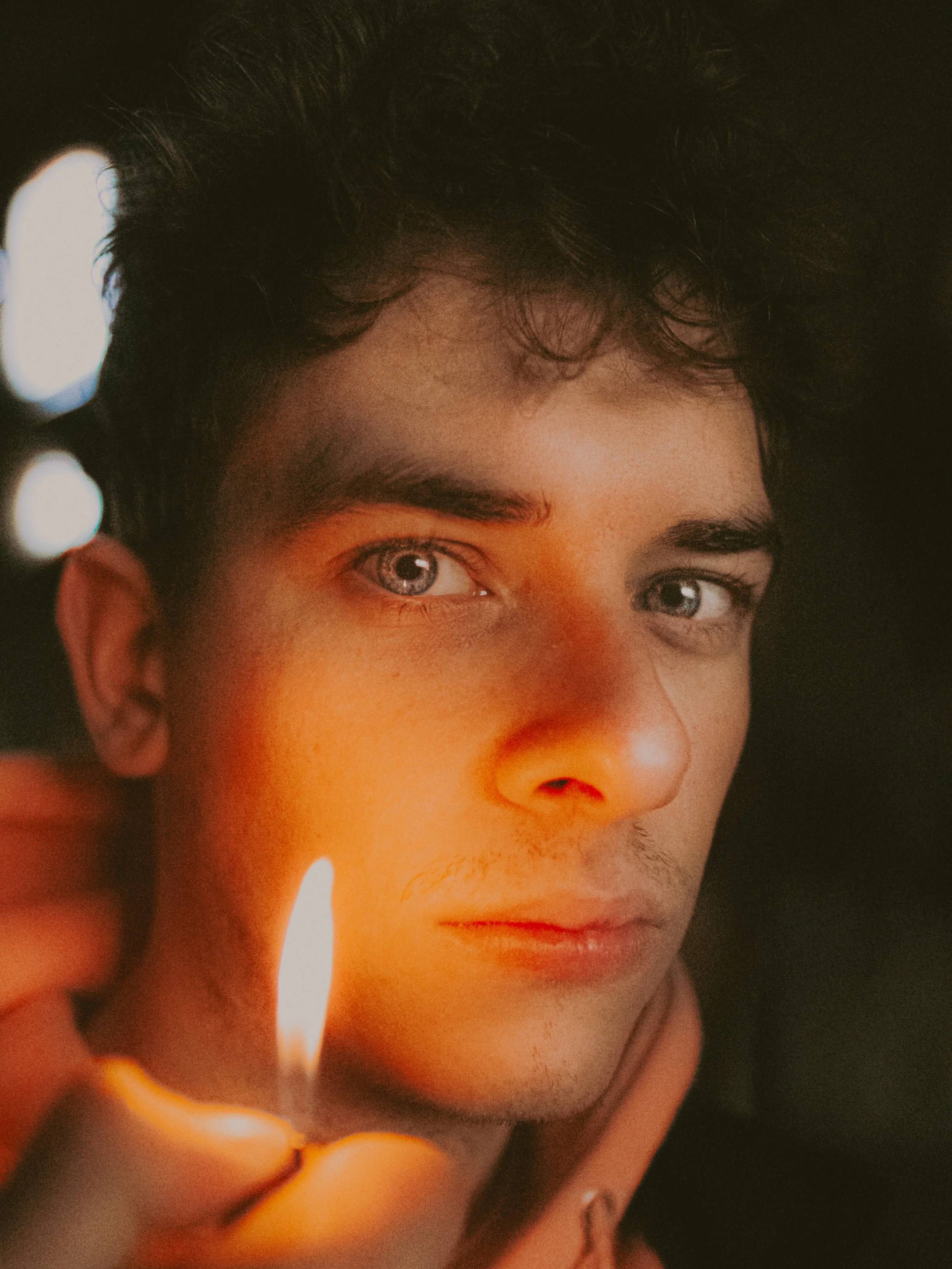 Jeune homme avec des yeux bleus, tenant une flamme devant son visage dans un environnement sombre.