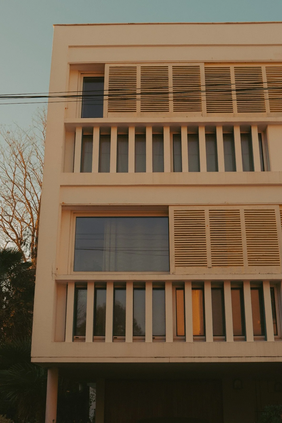 Façade d'un immeuble moderne avec plusieurs fenêtres, certains avec des volets en lattes horizontales, et des barreaux verticales séparant les fenêtres. Le bâtiment est de couleur claire et la photo est prise lors d'une fin d'après-midi avec la lumiè