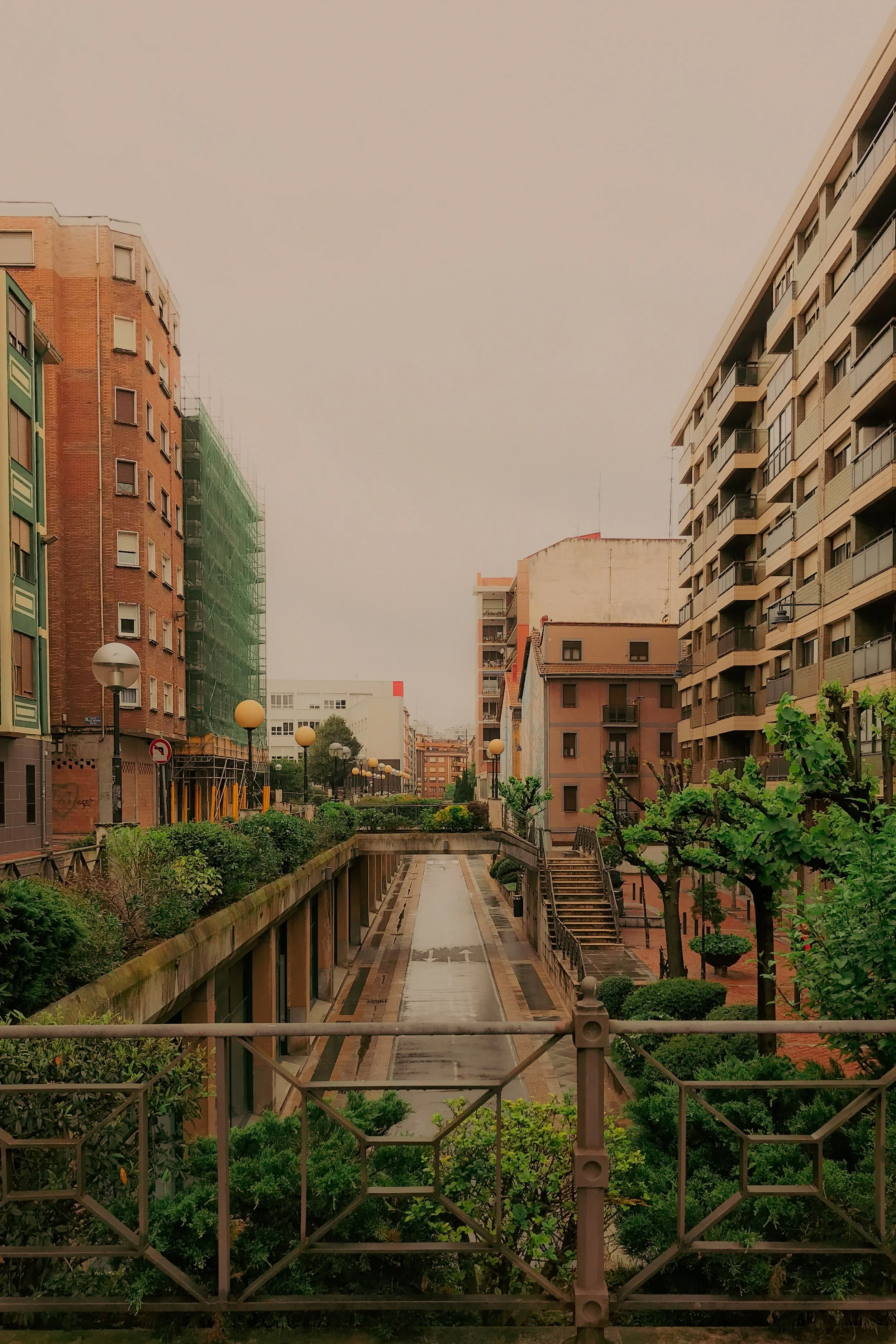 Vue d'une rue ou d'une allée urbaine bordée d'immeubles résidentiels et de lampadaires, avec des végétaux et un petit escalier en bois à droite.