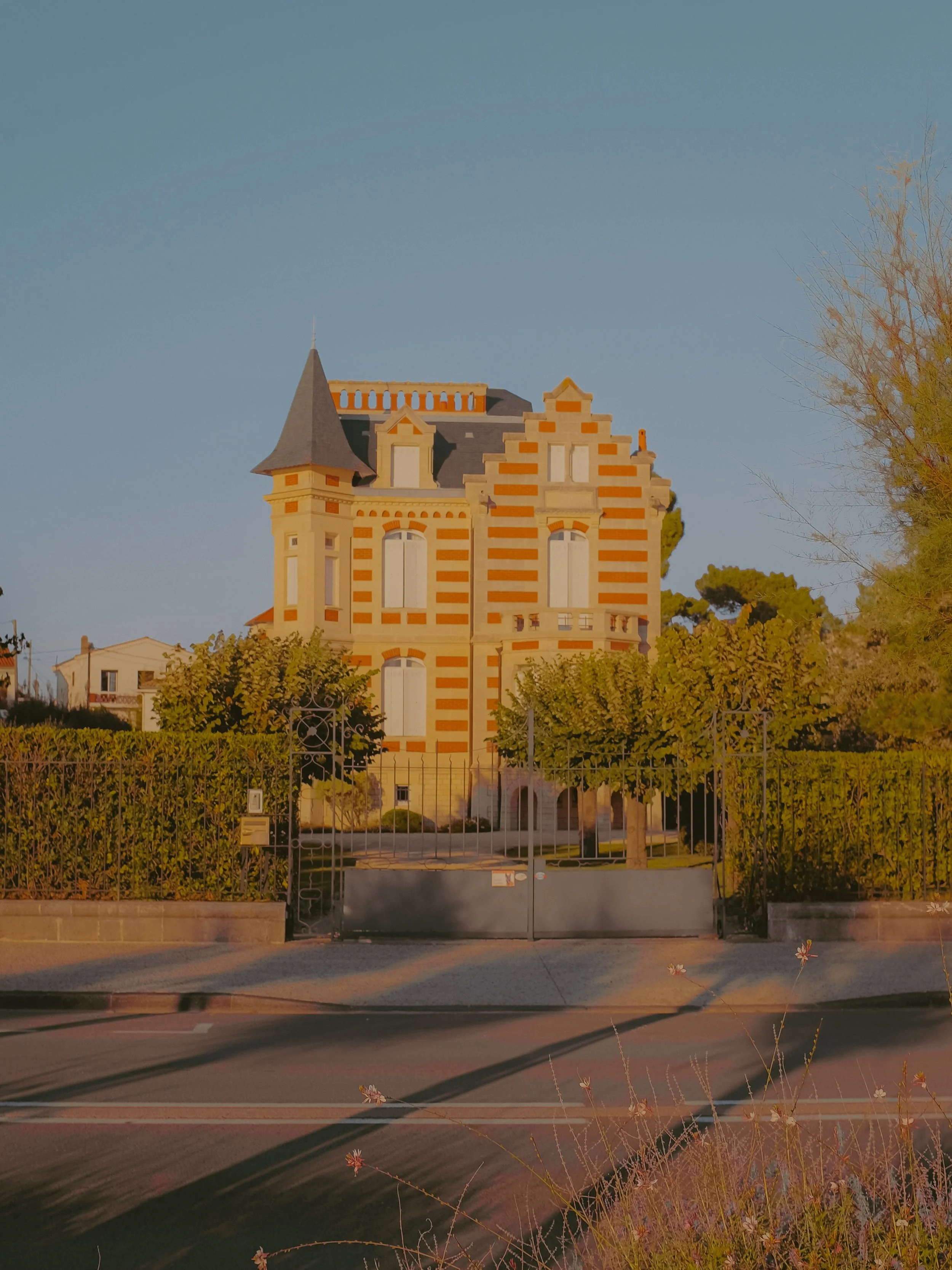 Une maison de style château avec une façade à rayures orange et beige, des fenêtres hautes, un toit pointu, entourée d'un portail en métal et de végétation verte, sous un ciel clair.