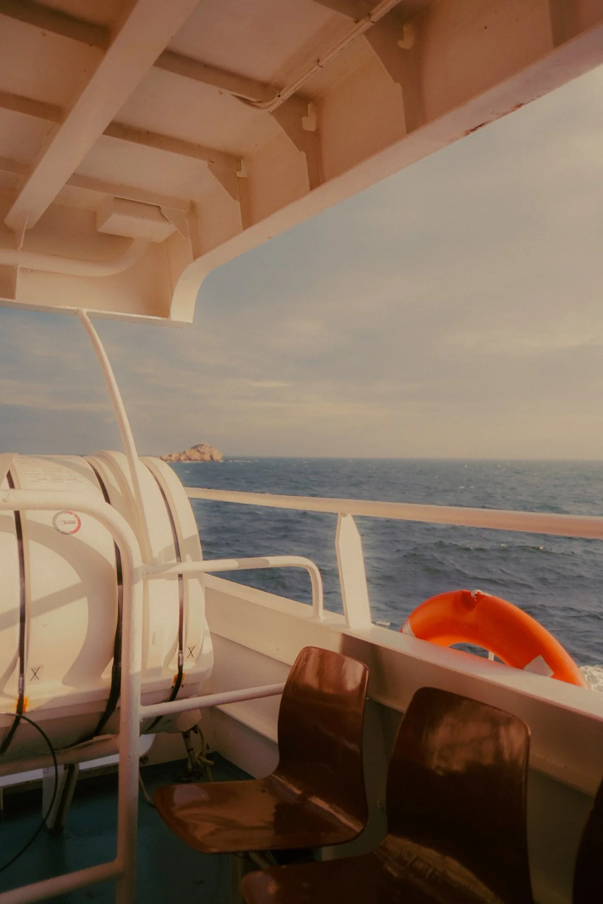 Vue depuis un bateau montrant la mer, un rocher au loin, une partie de la cabine avec des chaises en bois et une bouée orange sur la rambarde.