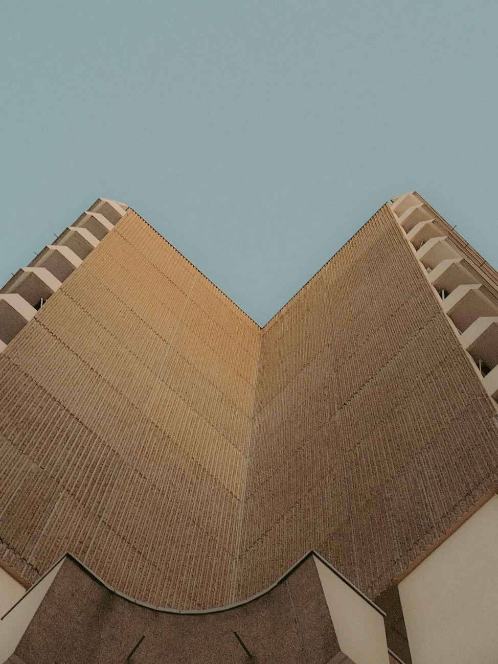 Vue d'un bâtiment en contre-plongée, montrant la face avant avec une texture en brique beige et un ciel bleu clair en arrière-plan.