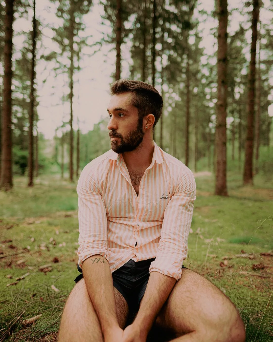 Un homme avec une barbe et des cheveux courts, portant une chemise à rayures roses et blanches, est assis dans une forêt avec des arbres autour, regardant vers la gauche.