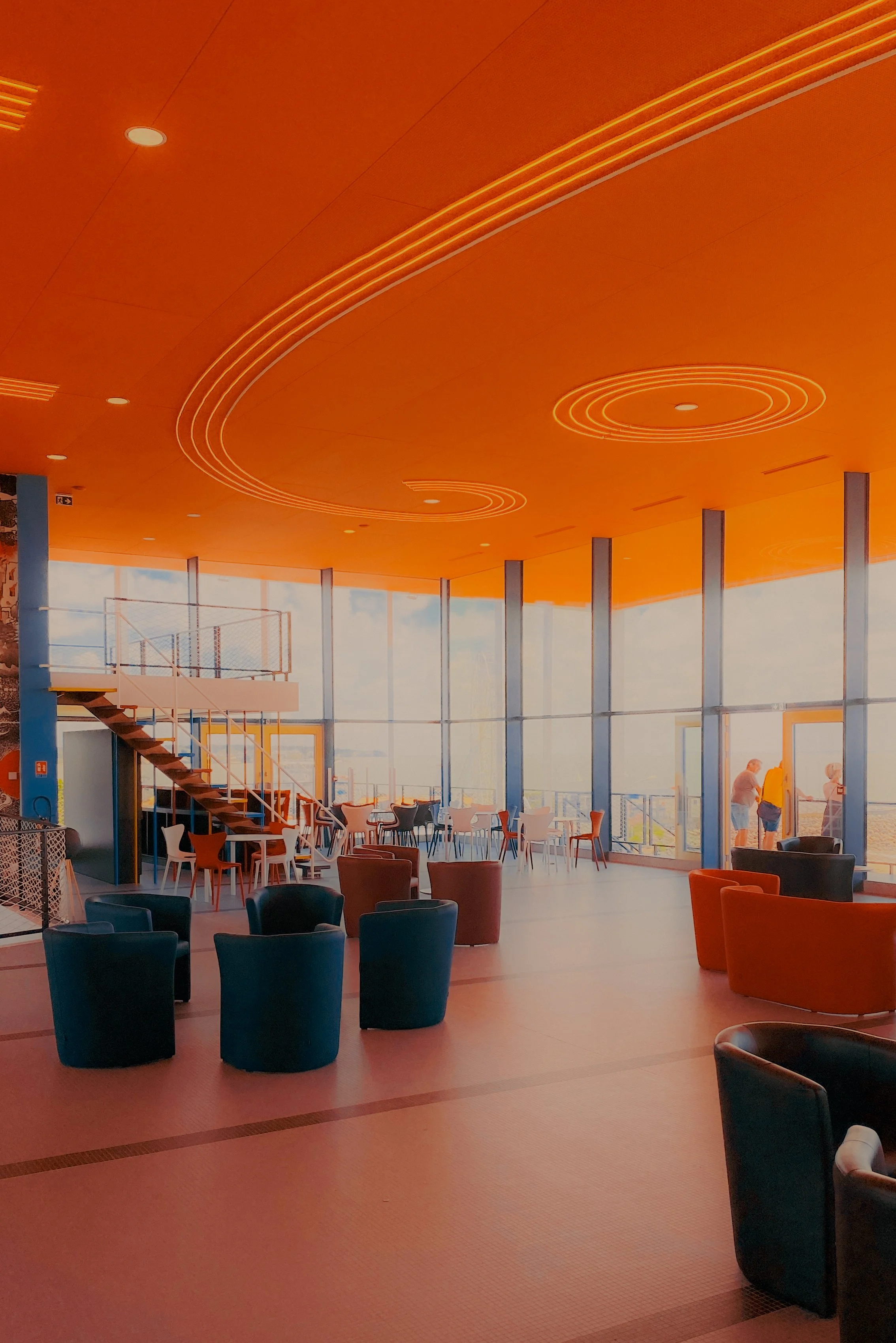 Un intérieur moderne avec de grandes fenêtres laissant entrer beaucoup de lumière naturelle, des fauteuils colorés disposés autour, une mezzanine avec des tables et chaises, et des personnes dehors visibles à travers les fenêtres.