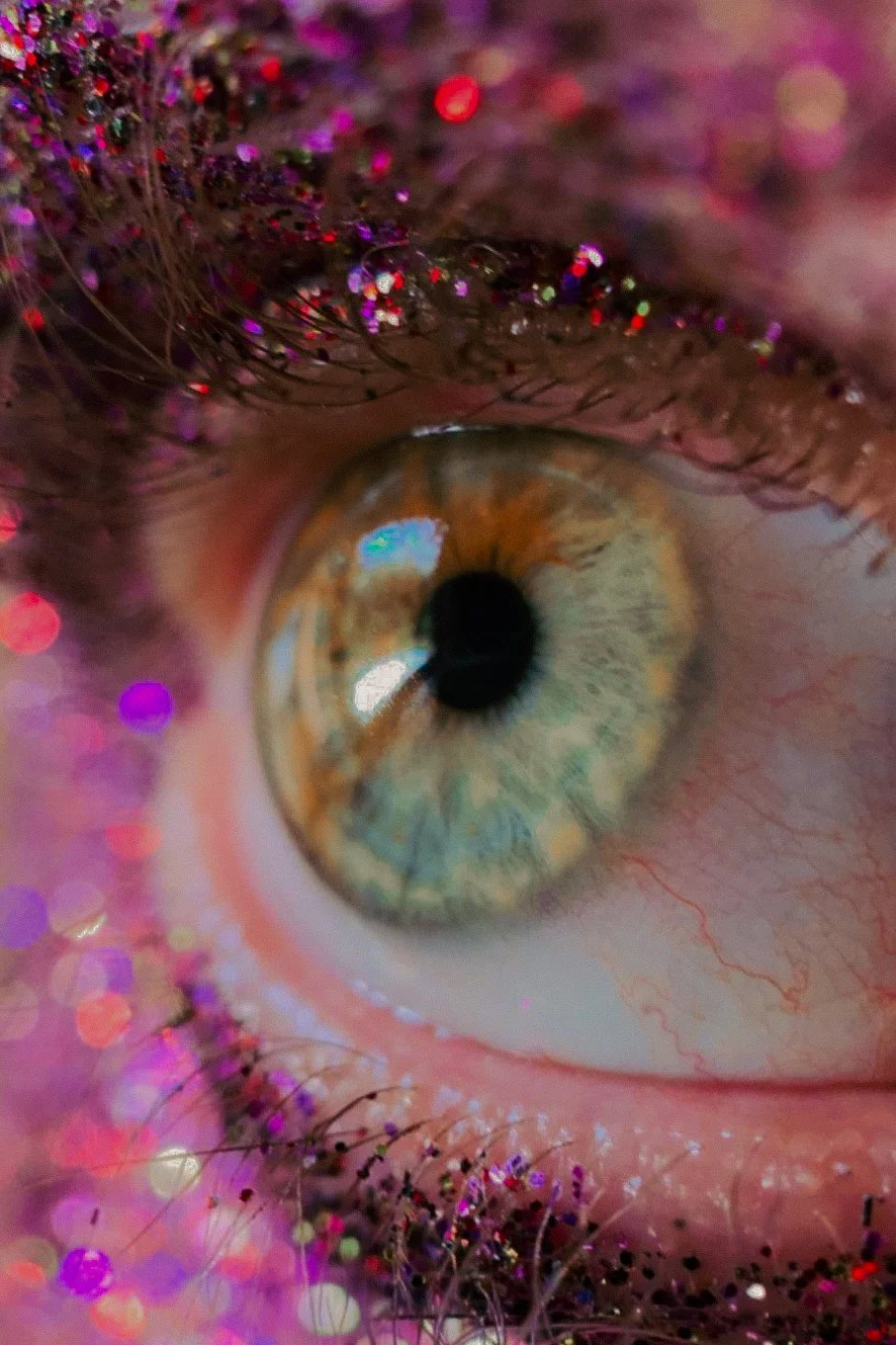 Un œil humain avec des reflets de lumière multicolores sur la pupille et les cils, entouré de paillettes colorées.