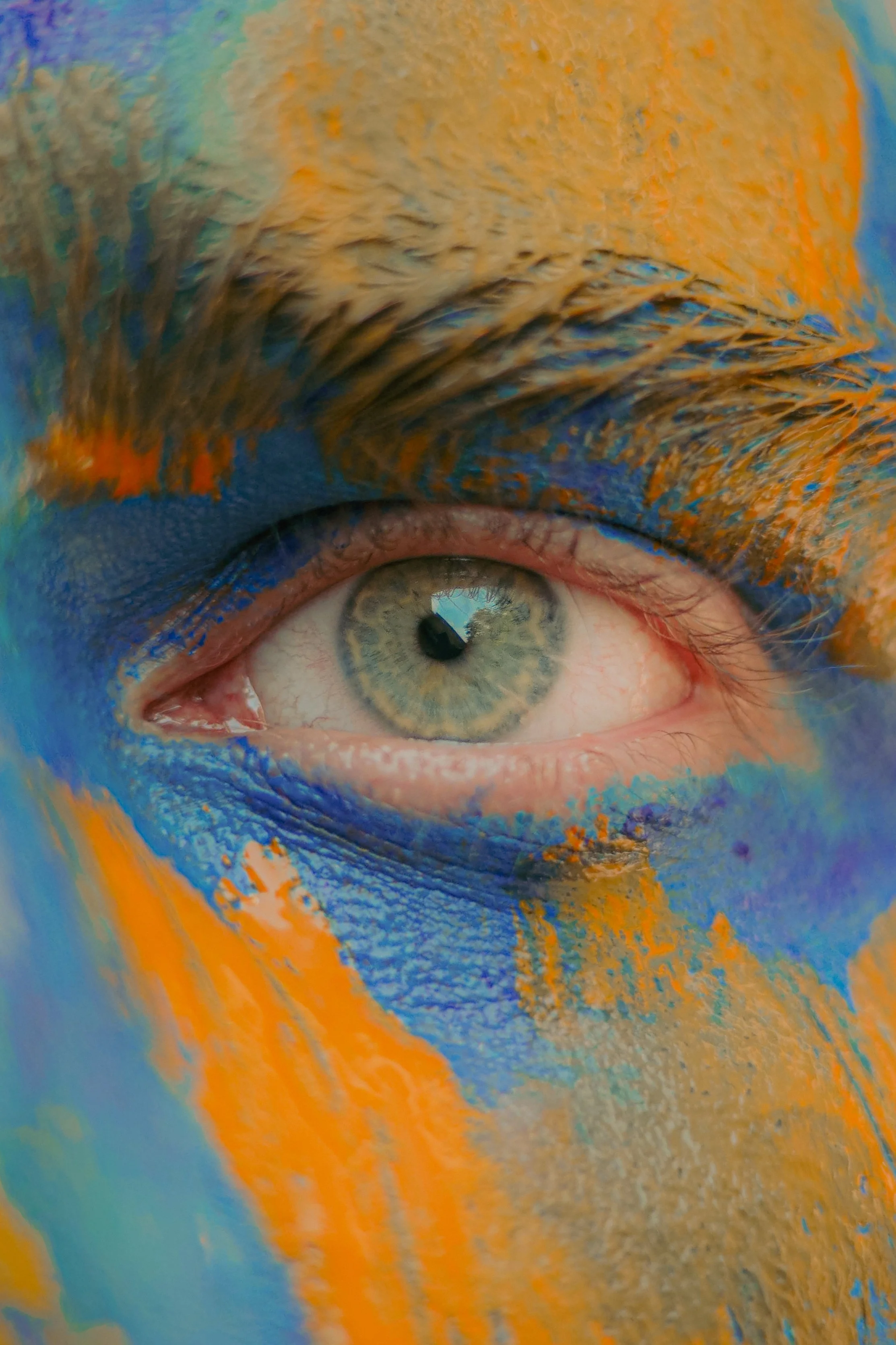 Un œil humain avec un maquillage coloré bleu, orange et jaune autour, peint sur le visage.