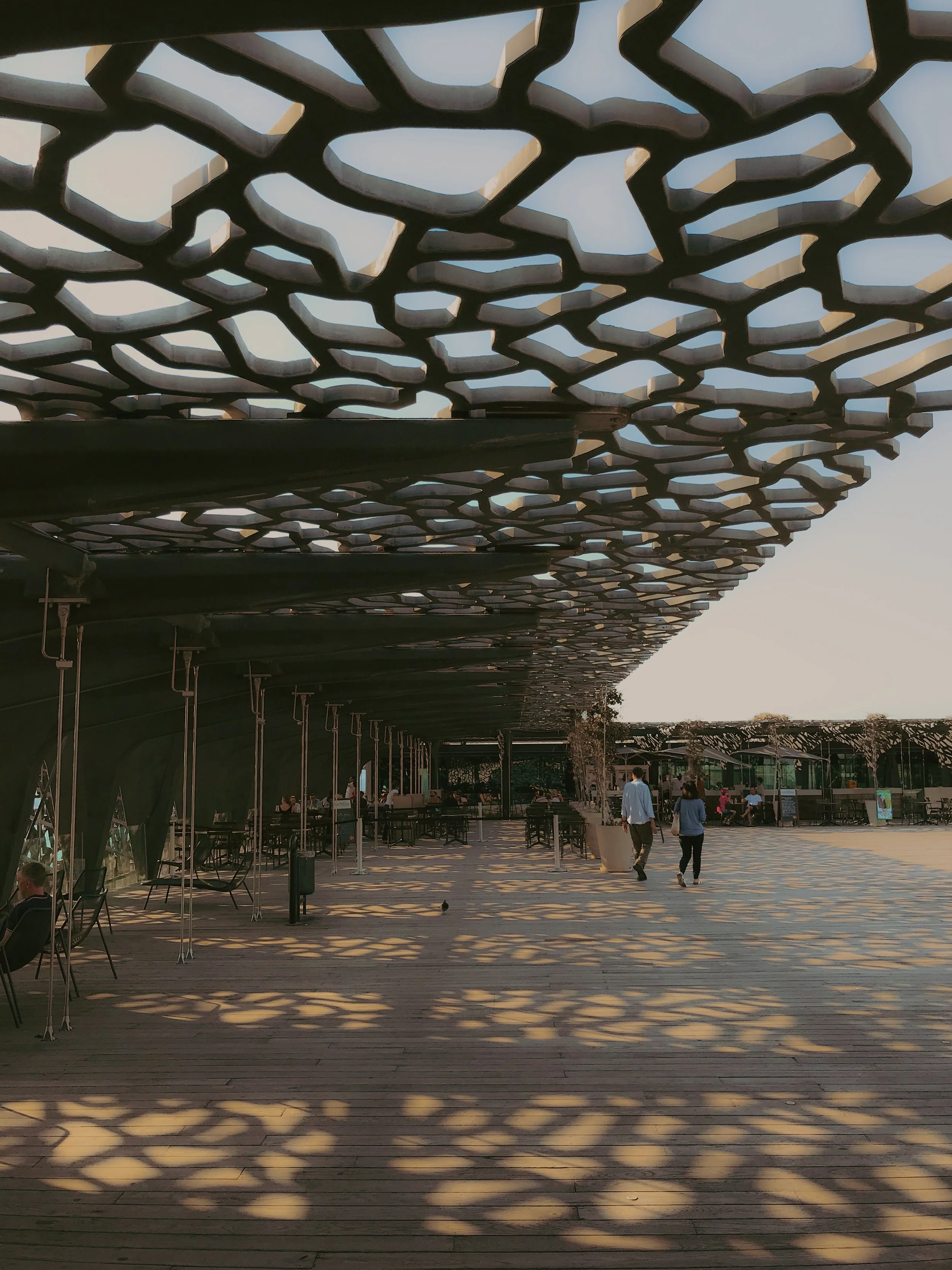 Une pergola en structure métallique avec un motif géométrique complexe qui crée des ombres décoratives sur le sol en bois. Au fond, on voit des personnes marchant, assises, et un espace public en plein air lors d'une journée ensoleillée.