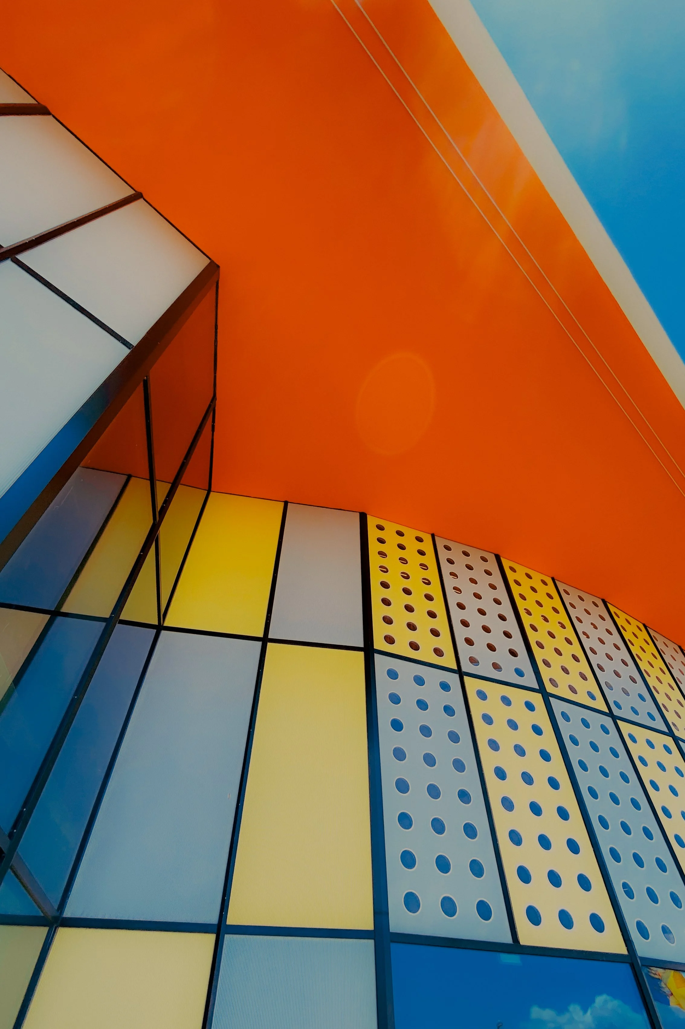 Façade d'un bâtiment avec des panneaux colorés jaune, blanc, et gris, certains avec des motifs de points bleus, et un mur orange au plafond, sous un ciel bleu avec quelques nuages.