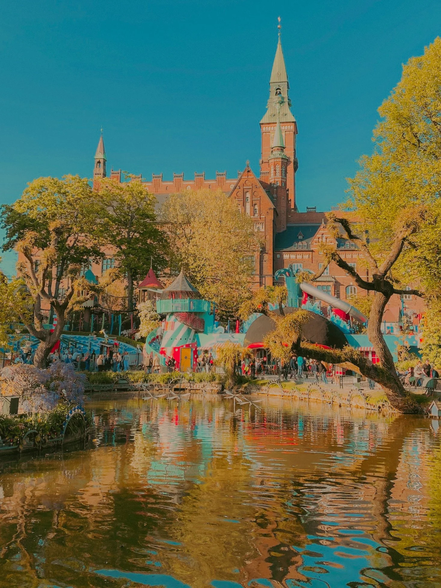 Un château en briques rouges avec des tours et un clocher, entouré d'arbres aux feuilles vertes, en face d'un étang reflétant le château et les arbres, avec des gens se promenant dans le parc.