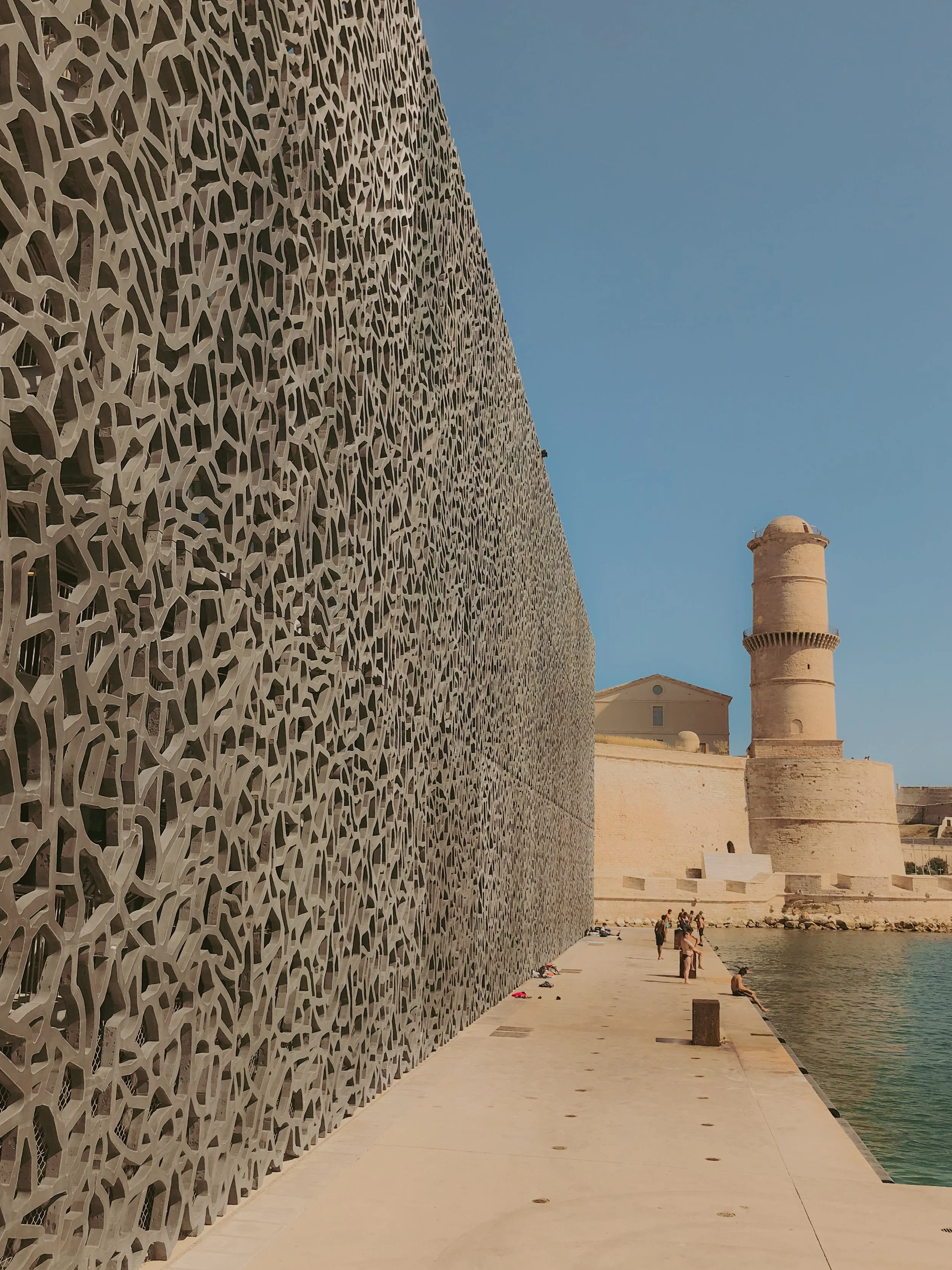 Une façade en pierre avec un rehaussement en métal avec un motif géométrique complexe. Au fond, un château ou une forteresse avec une tour ronde, située au bord de l'eau, sous un ciel bleu clair.