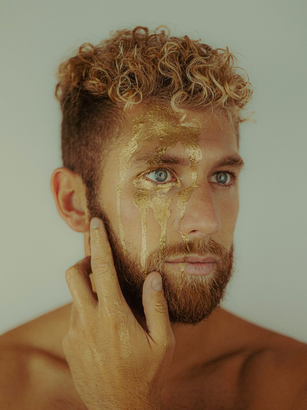 Un homme avec des cheveux bouclés blonds et une barbe brune, ayant de la peinture dorée ressemblant à de la peinture qui coule sur une partie de son visage, la main gauche touchant son visage.