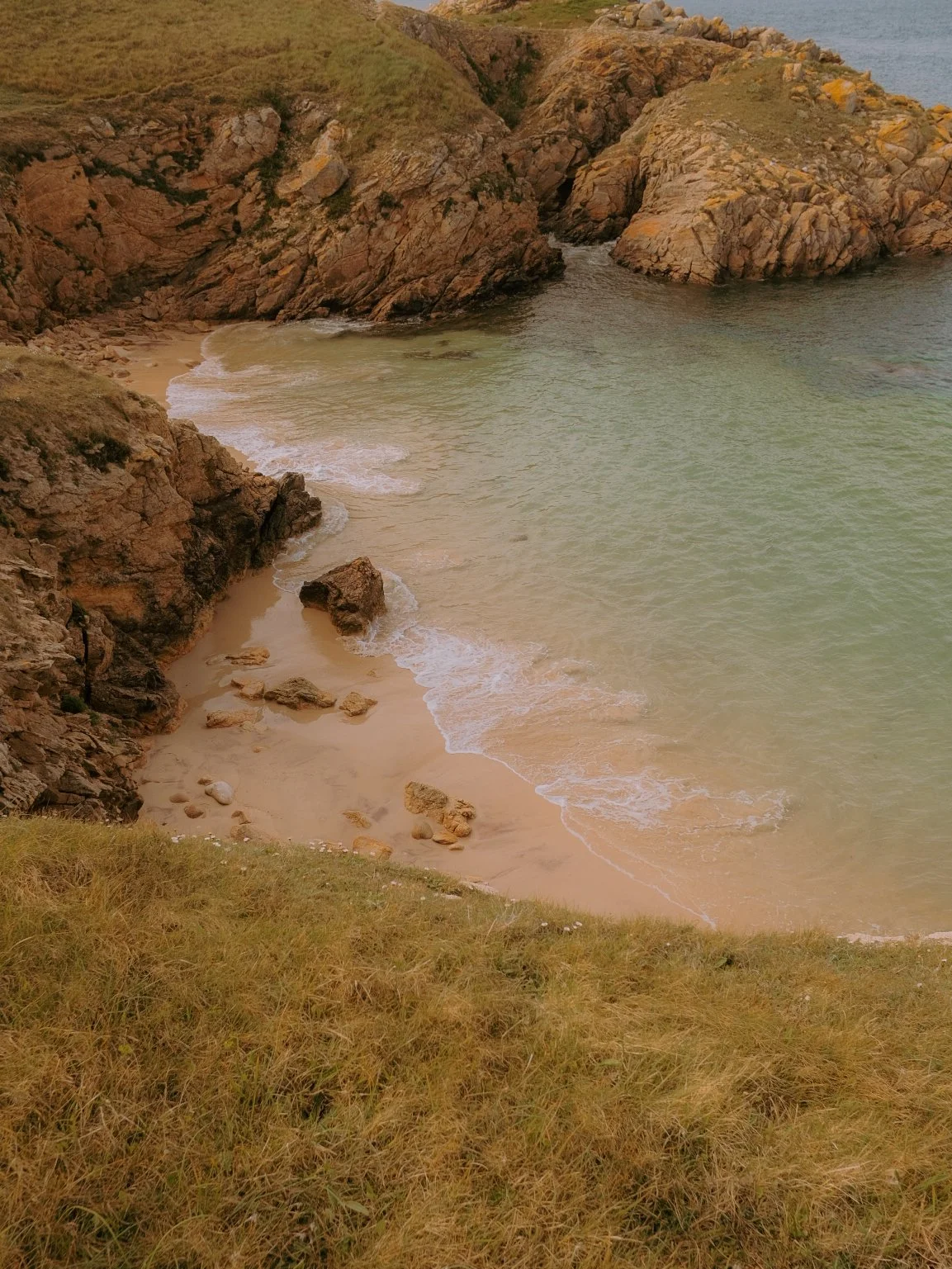 Plage isolée avec sable beige, rochers et falaises rocheuses vertes et brunes, vue de haut, ocean calme.