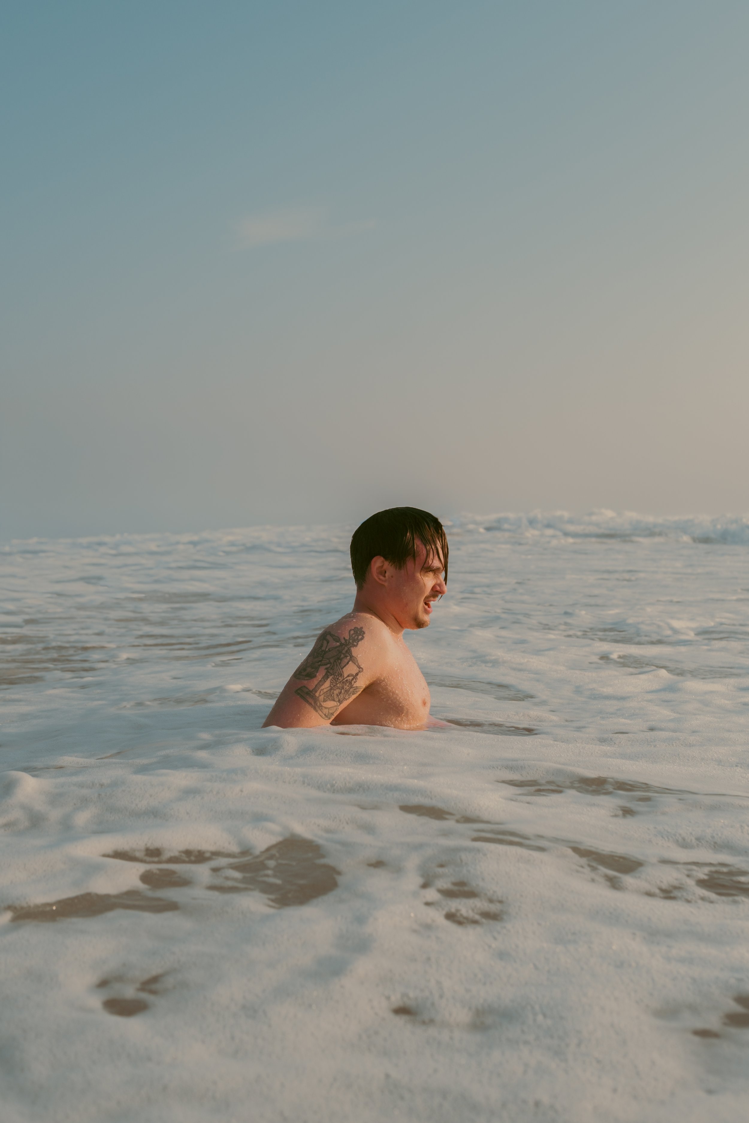 Un homme nu dans l'océan, avec tatouages sur le bras, à la plage, à l'aube ou au crépuscule.