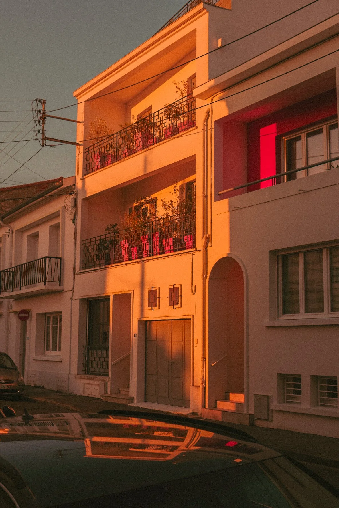 Une façade de bâtiment résidentiel blanc avec deux balcons en fer forgé noir, ornés de plantes, sous un coucher de soleil orange. La partie à droite est encadrée par une ombre qui crée un contraste chaud.