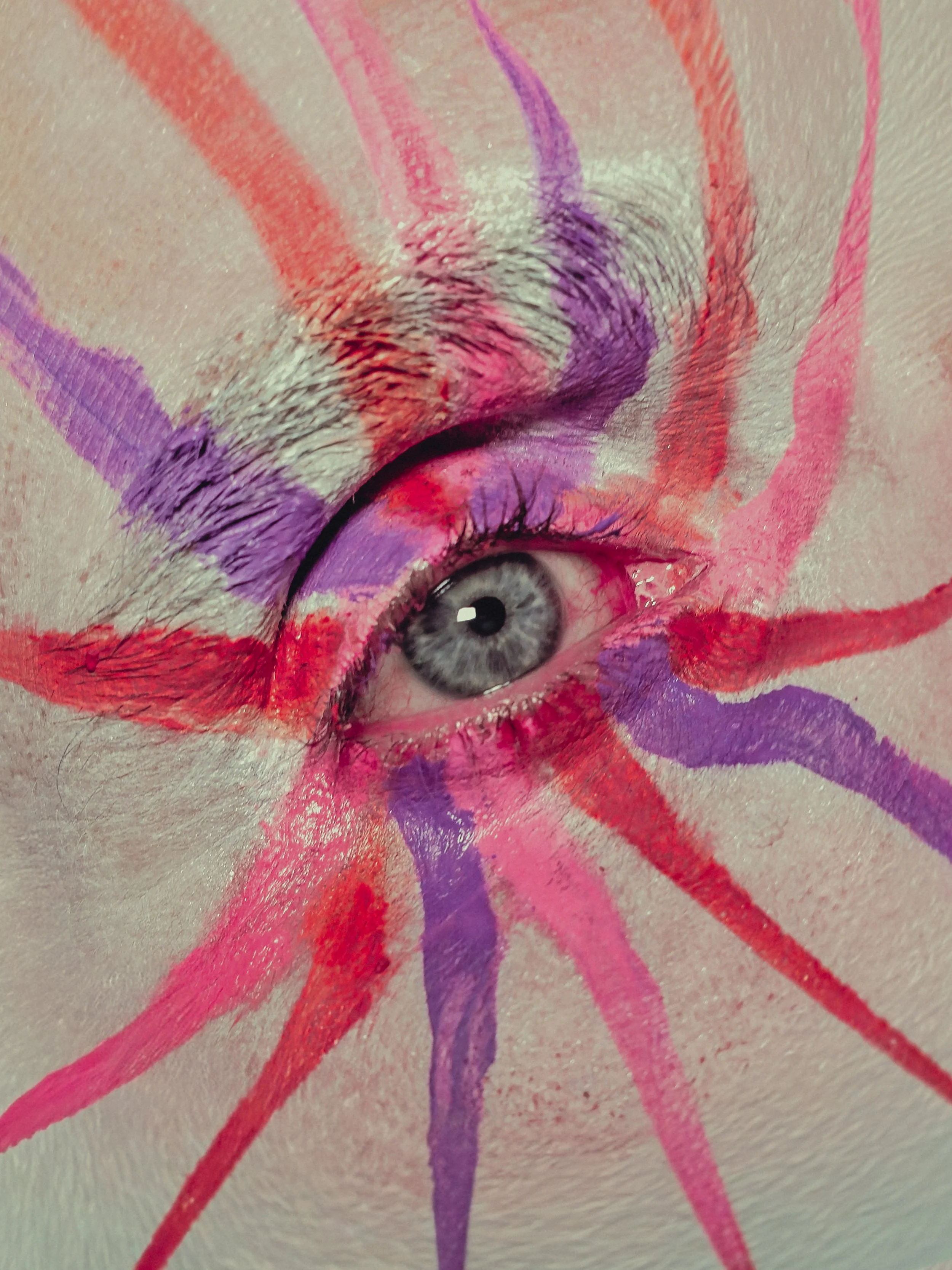 Un œil gris entouré de griffures peintes en rouge, violet, rose et orange sur le visage.