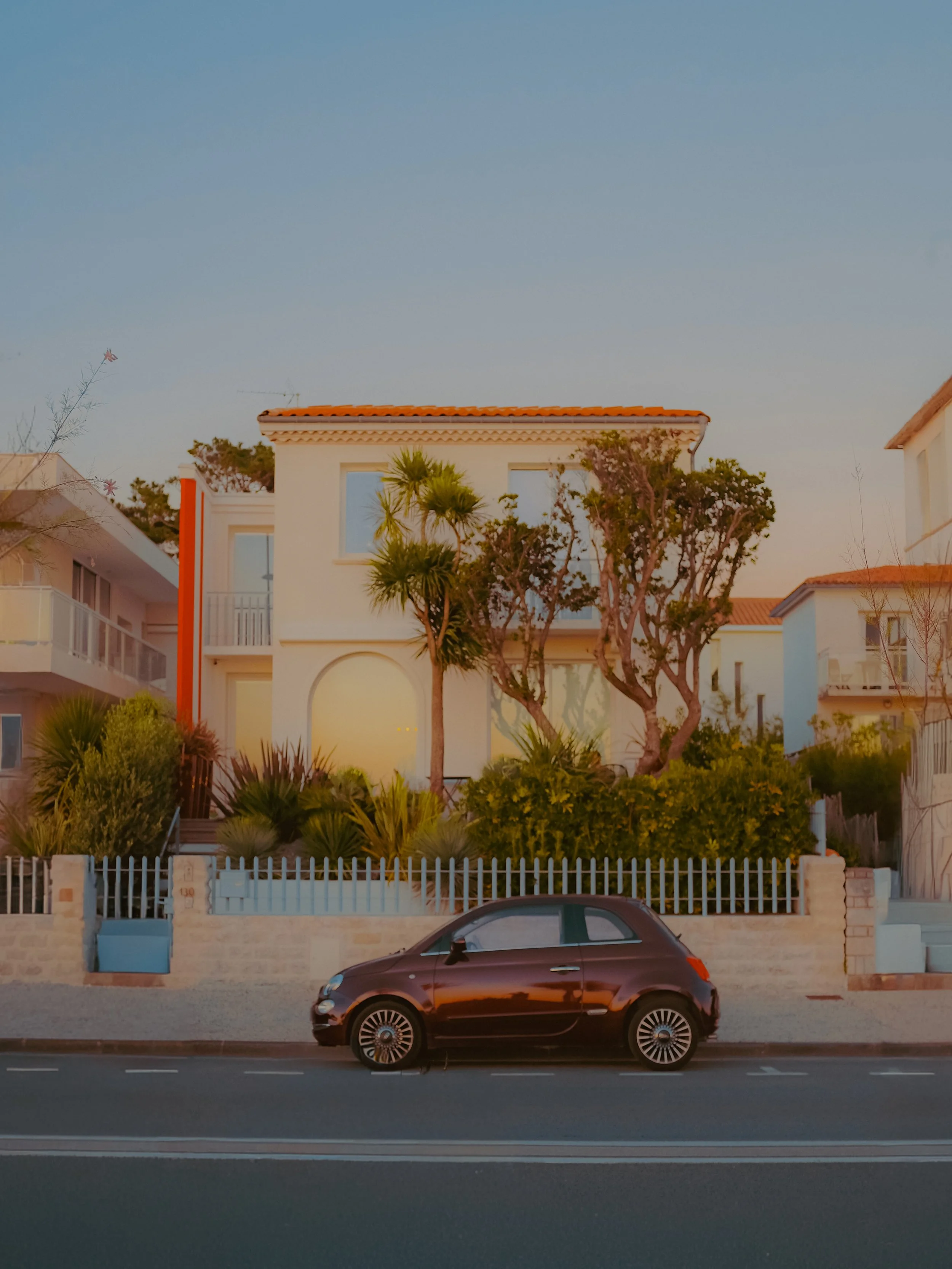 Une voiture noire garée devant une maison blanche, avec un jardin verdoyant et un ciel clair. 