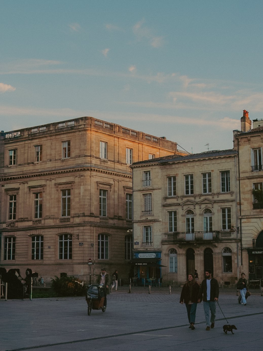 Vue de bâtiments historiques avec des fenêtres et une terrasse en pierre, avec des personnes marchant dans une place pavée au coucher du soleil.