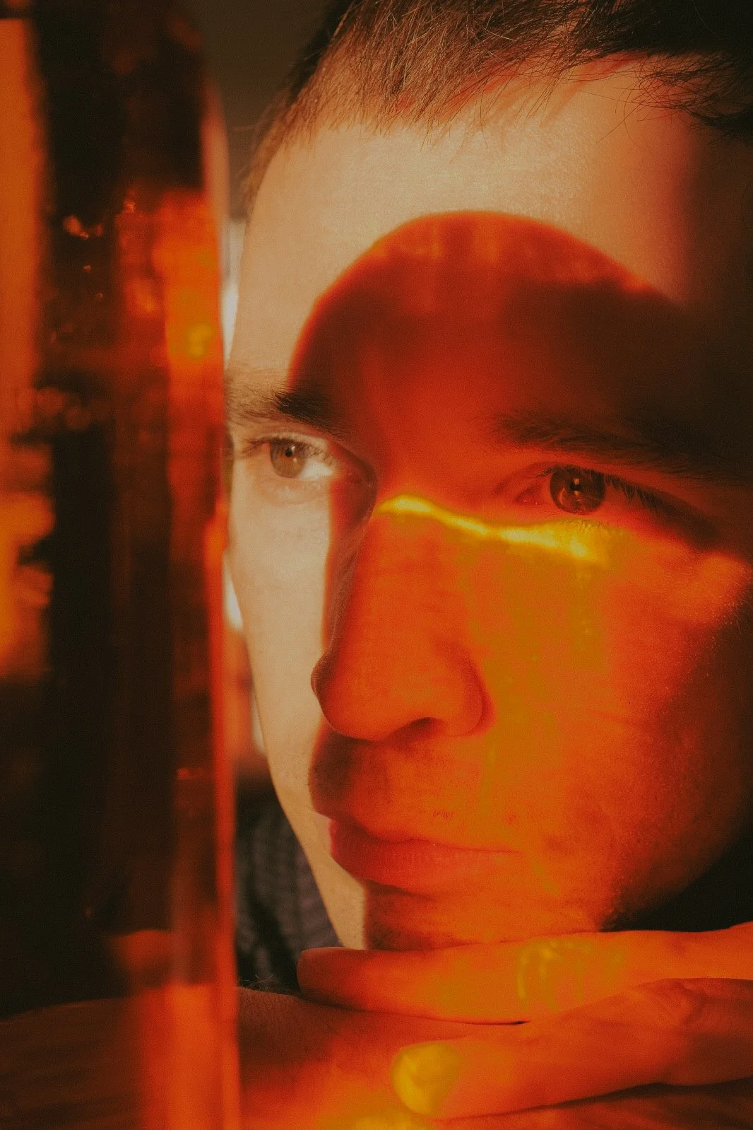 Portrait d'un jeune homme regardant pensivement, avec un effet de reflet ou de projection orange et rouge sur son visage.
