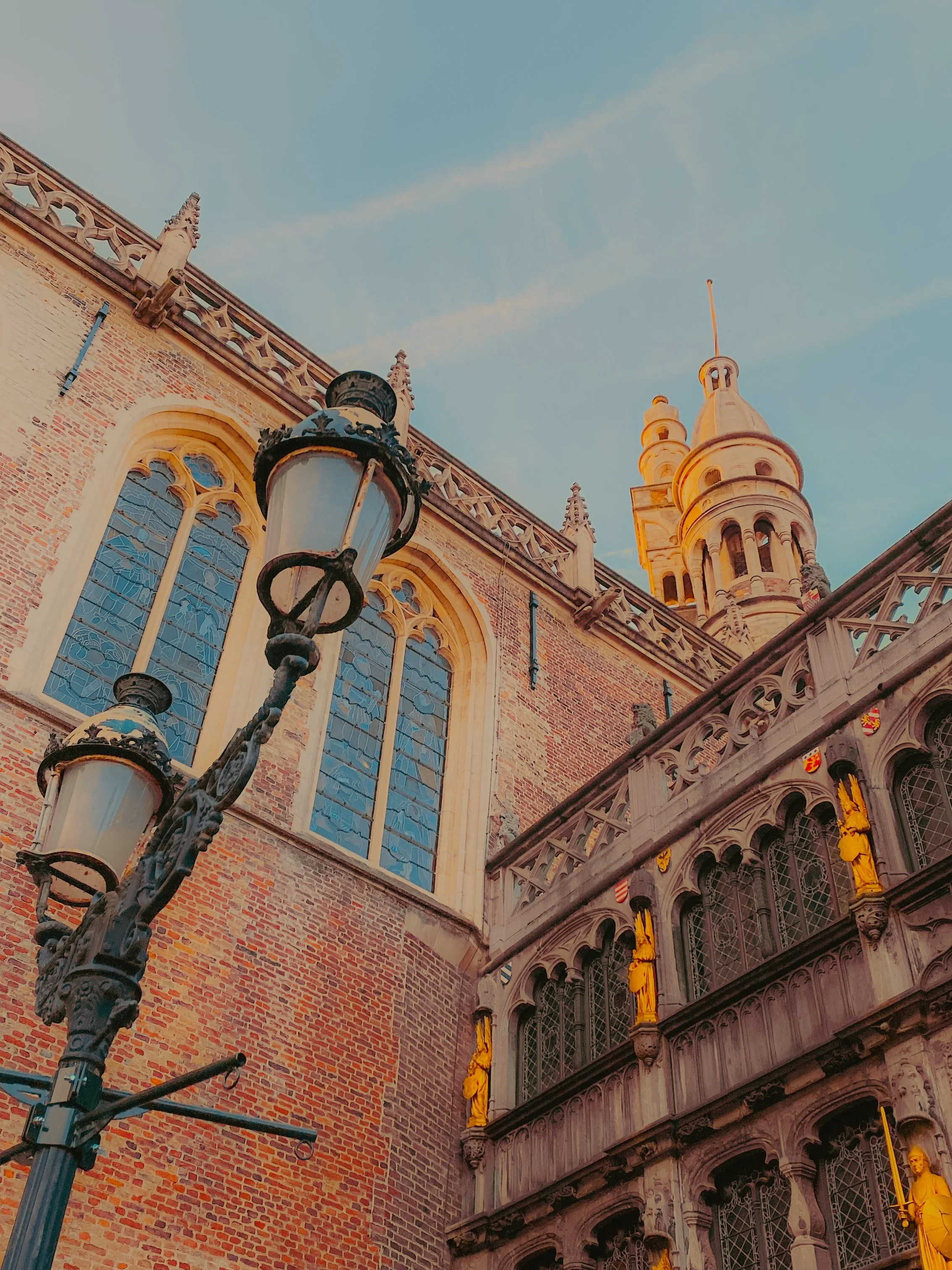Un bâtiment gothique avec de hautes fenêtres en verre coloré, des statues dorées sur la façade, et un lampadaire ancien au premier plan, sous un ciel bleu.