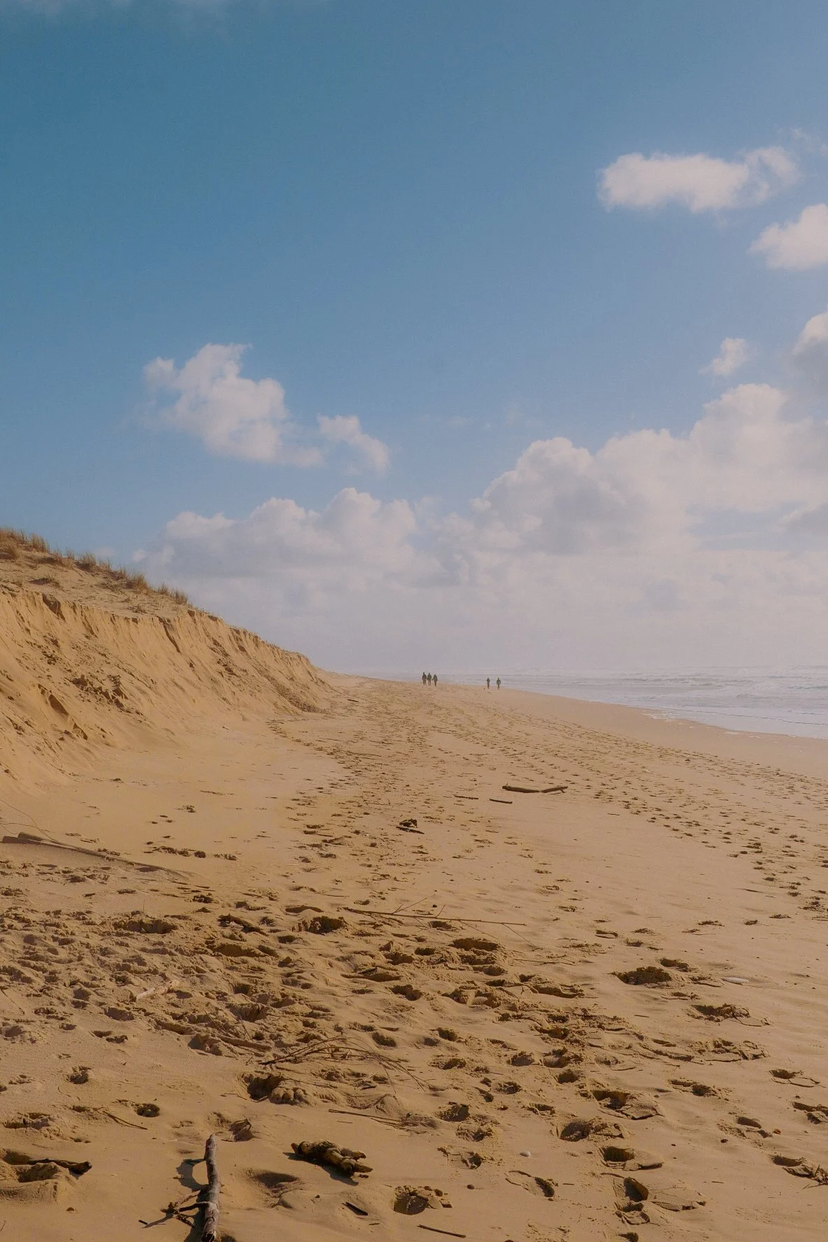 Plage de sable avec des dunes sur la gauche, ciel bleu avec quelques nuages et quelques personnes au loin marcher sur la plage.