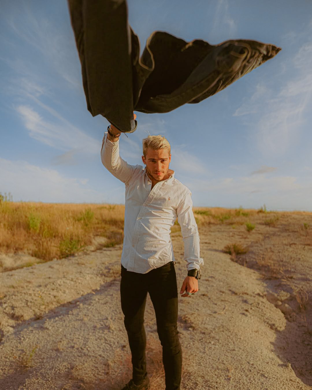 Un homme debout dans un champ désertique, portant une chemise blanche, seul, avec un ciel bleu en arrière-plan, tenant quelque chose dans sa main droite.