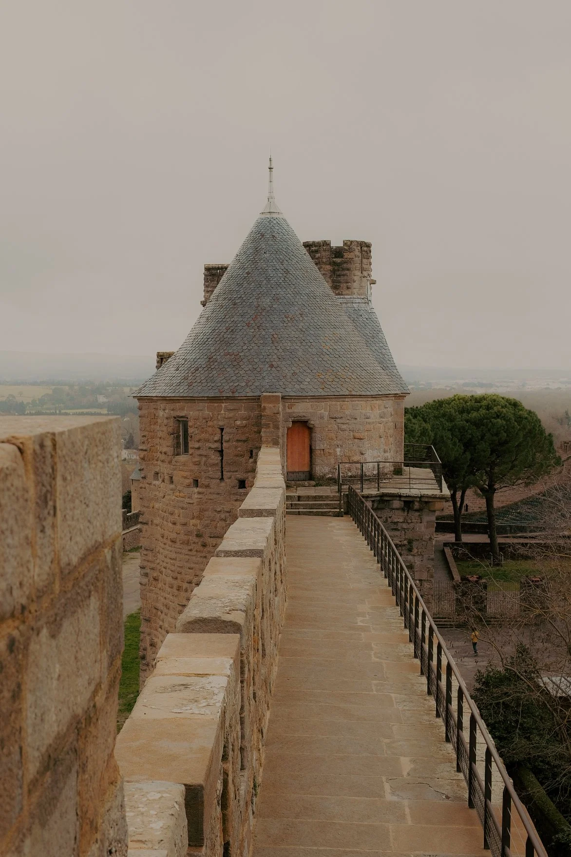 Tour d'enceinte médiévale en pierre avec une passerelle menant à une porte en bois, surmontée d'un toit conique en ardoise, vue d'un château, paysage rural en arrière-plan.