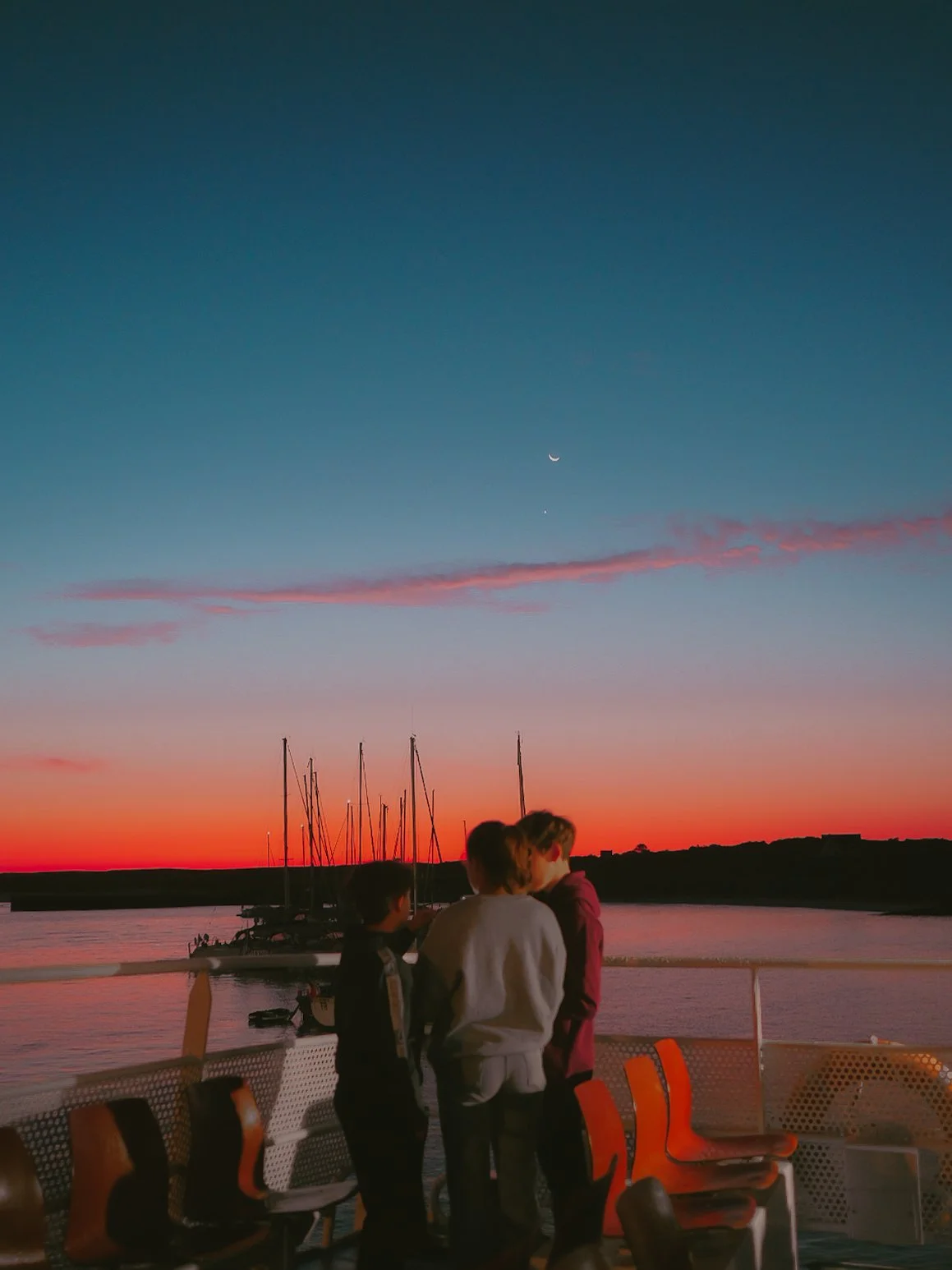 Trois personnes regardent le coucher de soleil sur une rivière depuis un bateau, avec un ciel coloré et la lune visible.