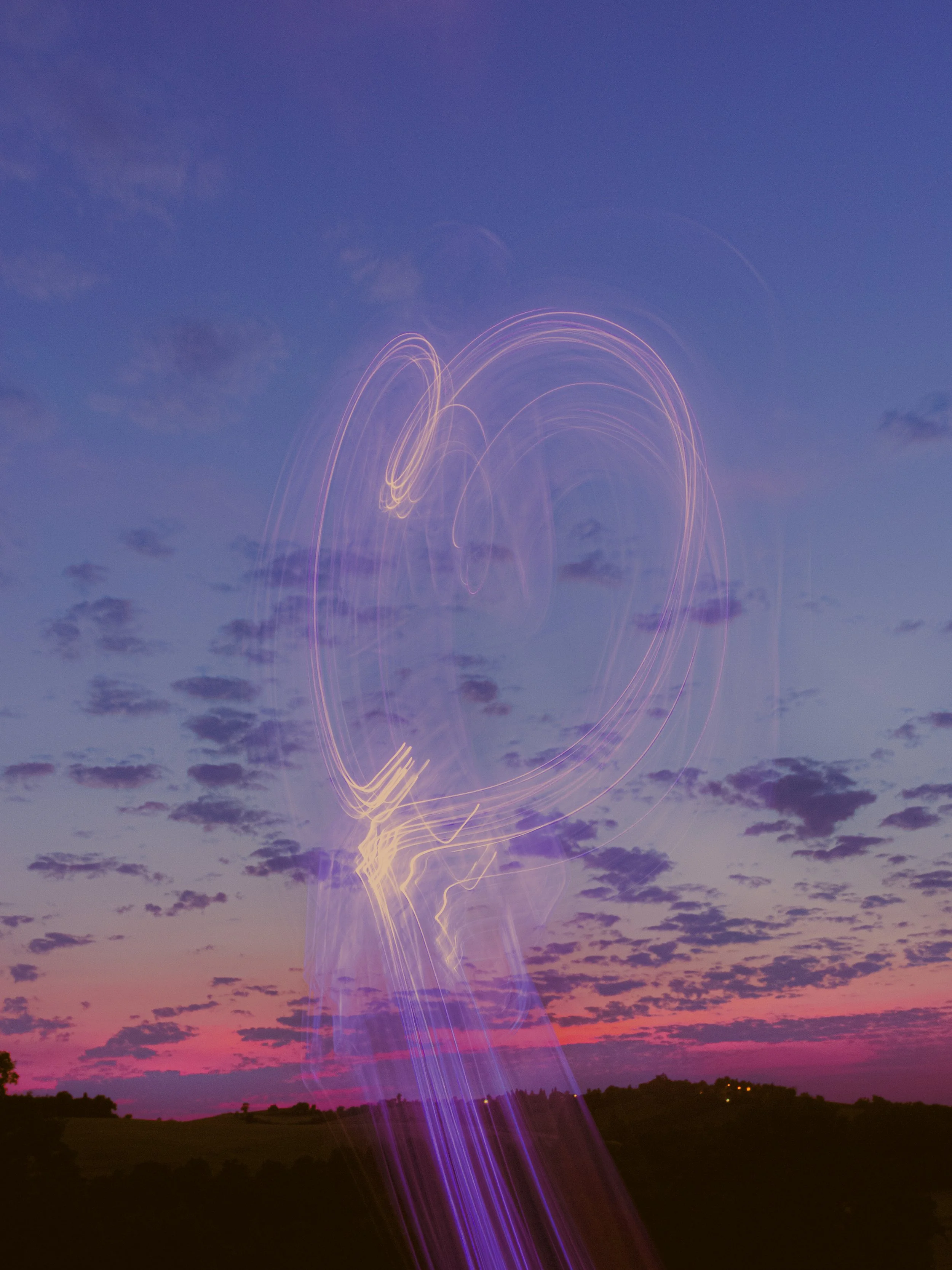 Ciel crépusculaire avec des nuages et des traînées lumineuses en forme de cœur dans le ciel, probablement causées par des feux d'artifice ou des lumières en mouvement.