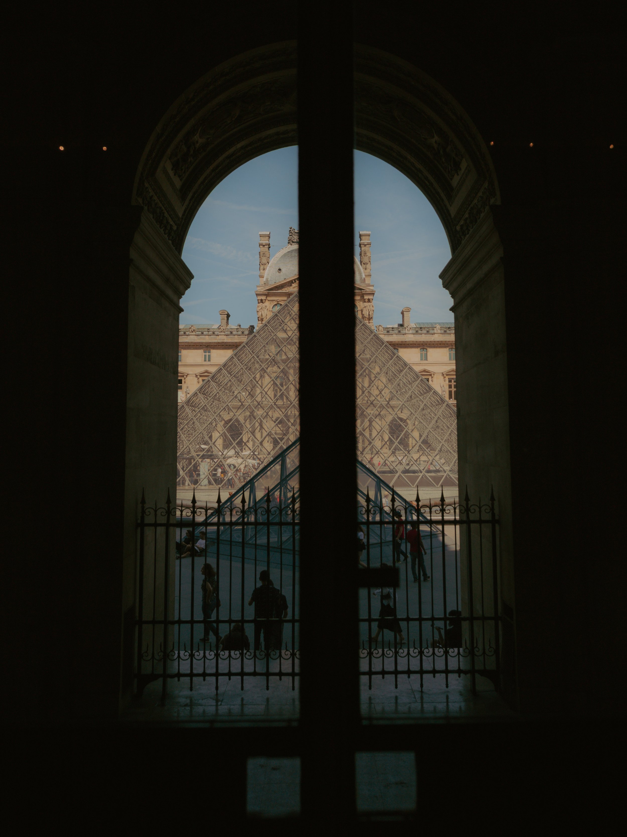 Vue de la pyramide du Louvre à travers une fenêtre, avec des visiteurs en bas et un bâtiment historique en arrière-plan.