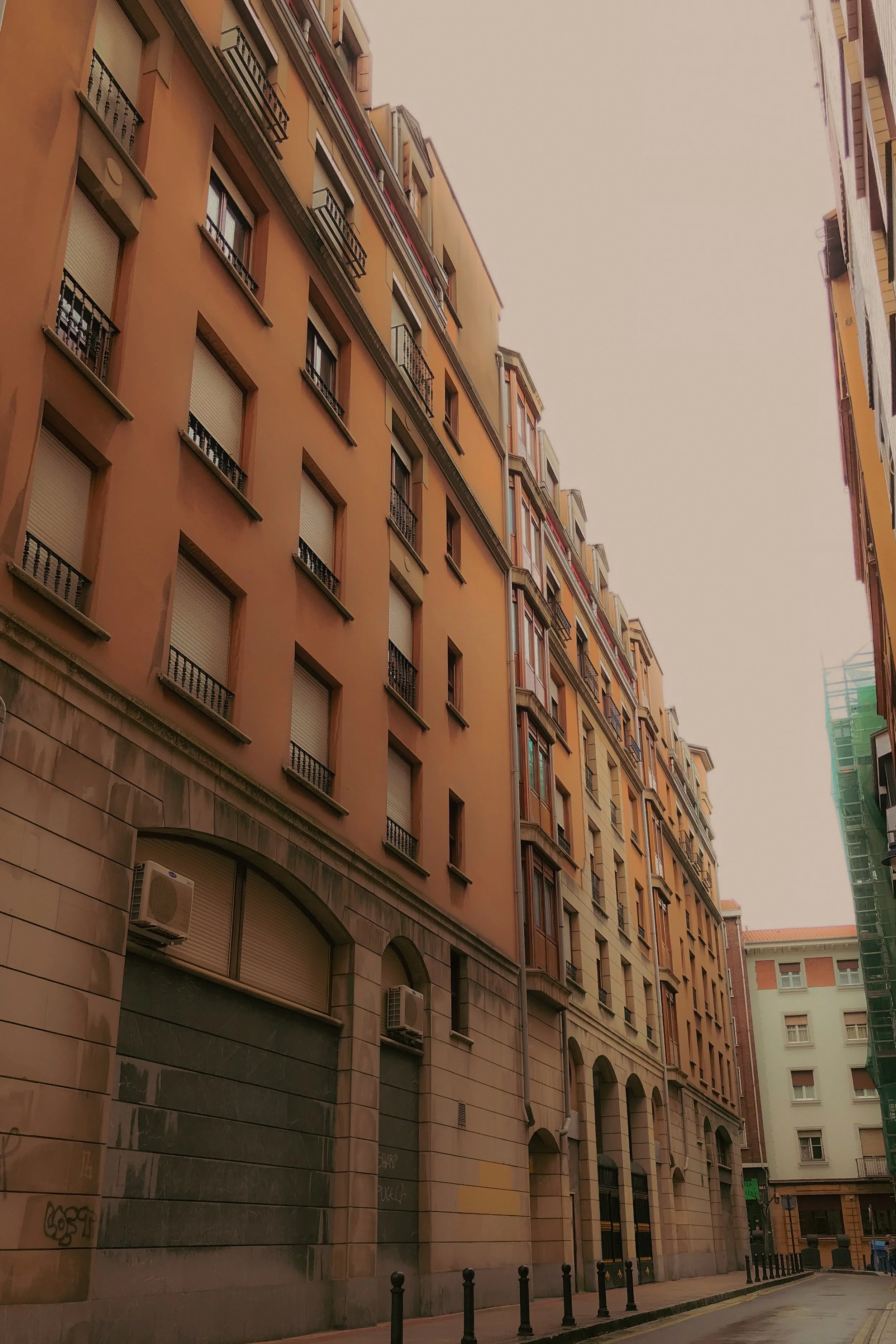 Immeubles résidentiels avec plusieurs étages, fenêtres avec volets roulants, petite rue calme, piétons et barrières de sécurité sur le trottoir.