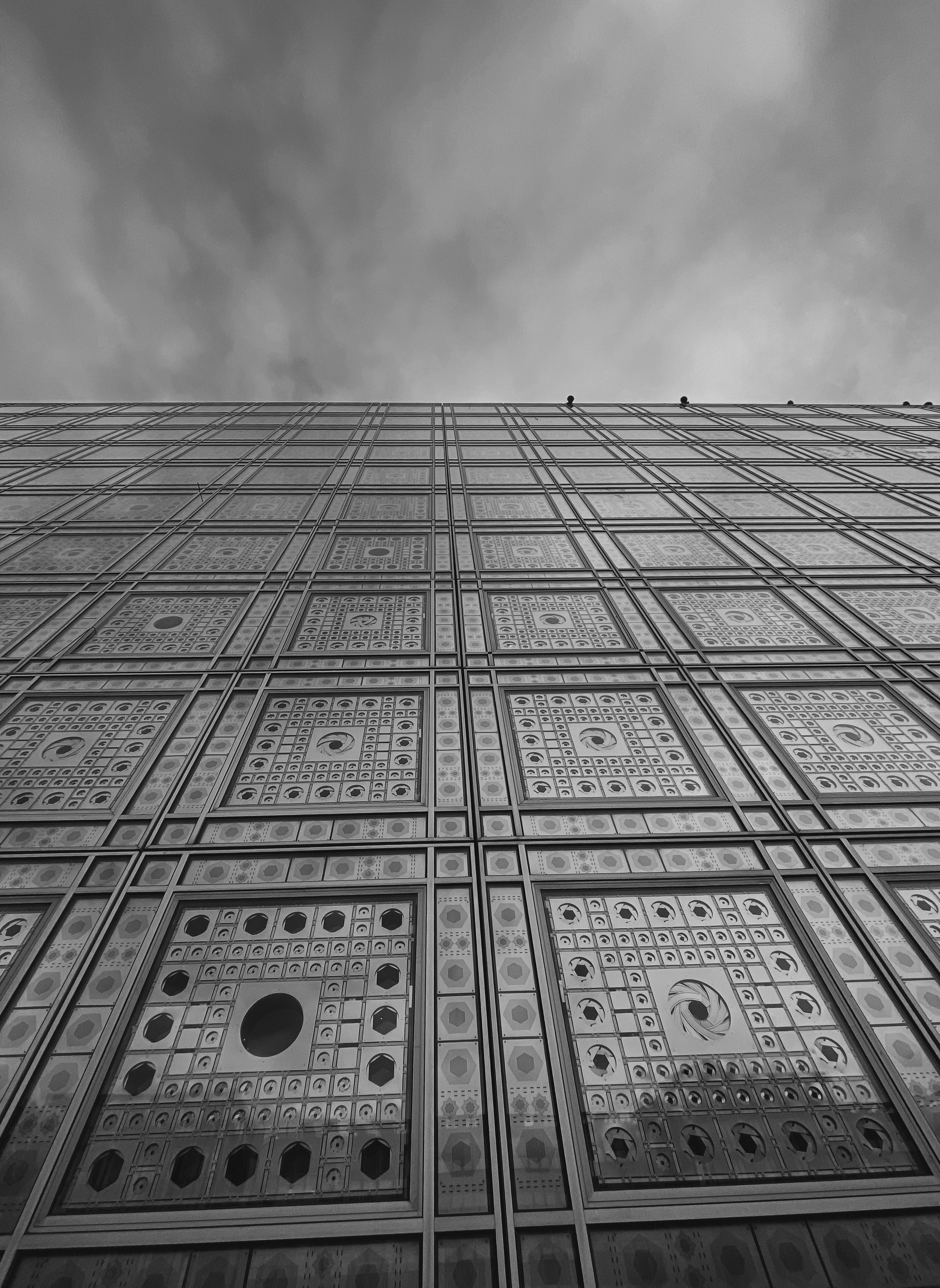 Façade d'un bâtiment avec une structure en verre ornée de motifs géométriques, vu de bas en haut sous un ciel nuageux.