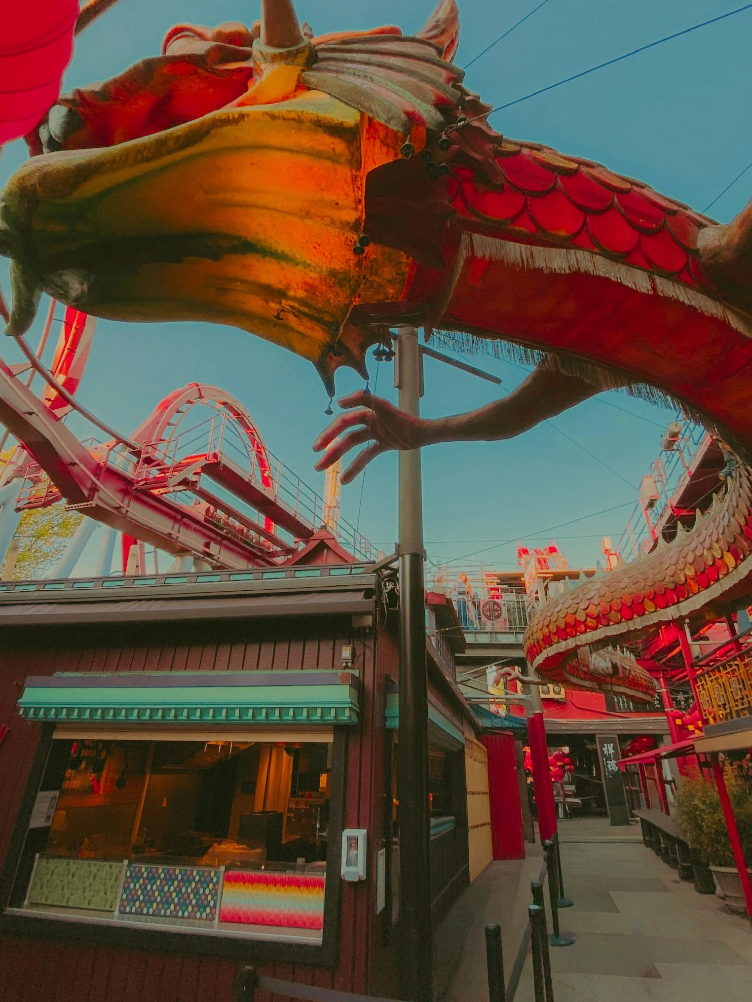 Décor de fête foraine avec une grande sculpture de dragon en haut, une structure de roller coaster en arrière-plan, et des bâtiments colorés en bas, sous un ciel clair.