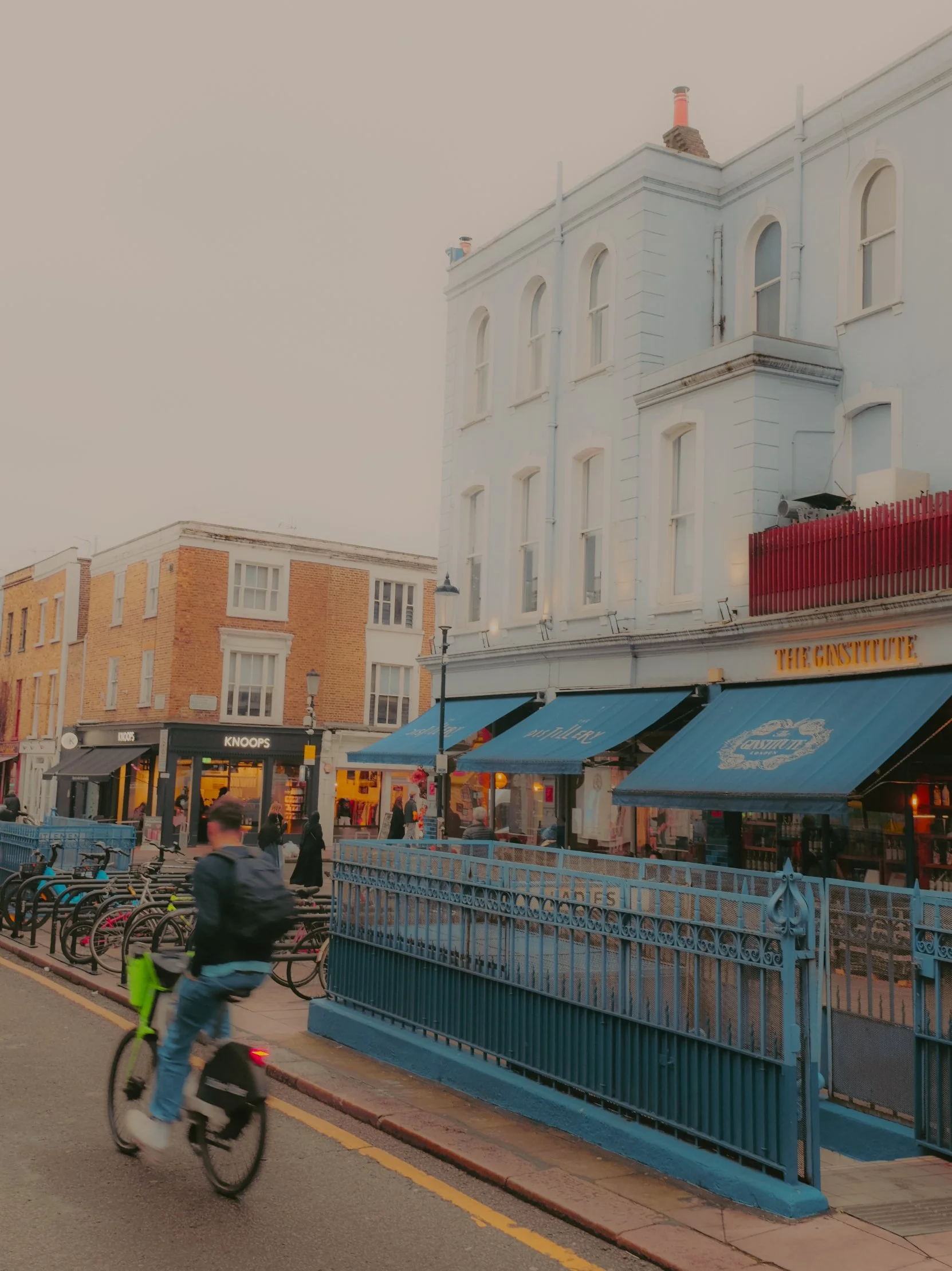 Rue urbaine avec un cycliste, des magasins avec enseignes, un bâtiment blanc avec des fenêtres et des auvents bleus.