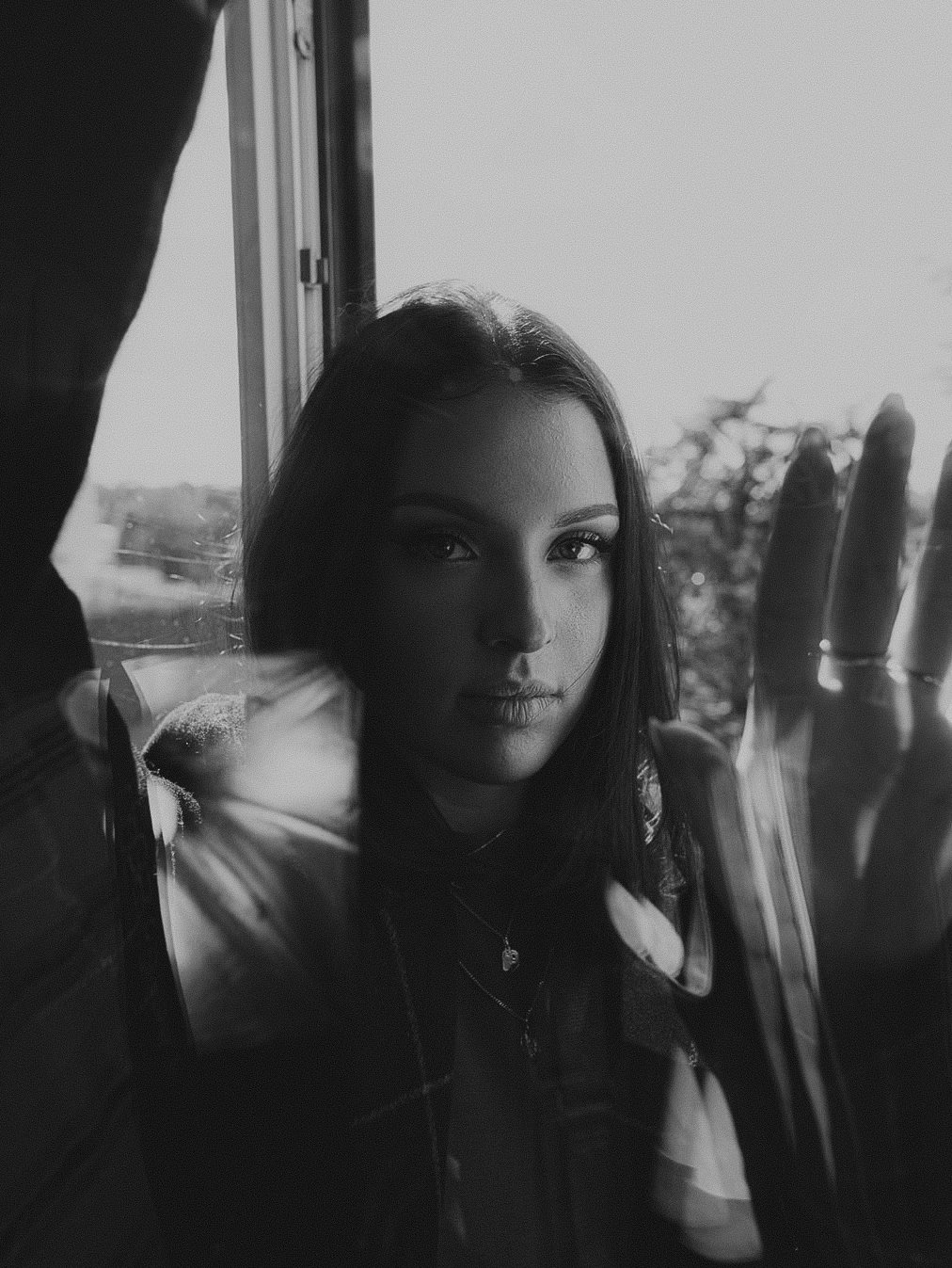 Une femme regarde par une fenêtre, de profil, avec la main contre la vitre. La photo est en noir et blanc.