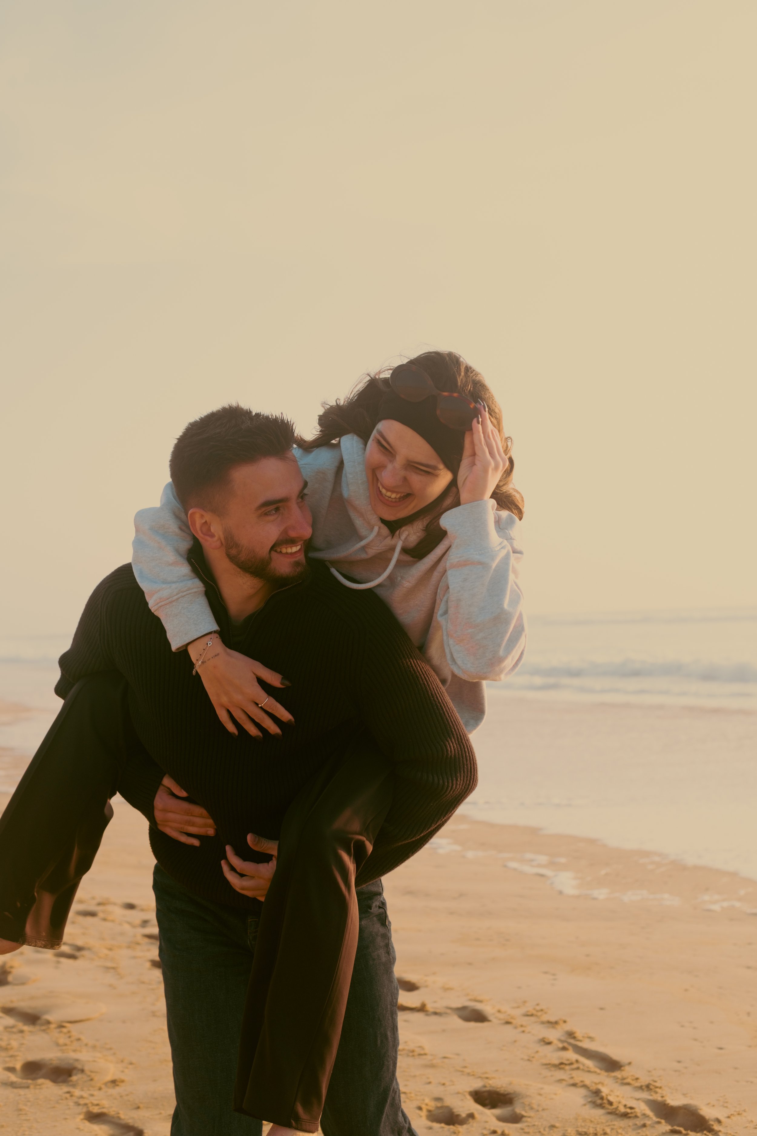 Un jeune couple souriant sur la plage au coucher du soleil, l'homme portant un sweat noir et la femme une sweat gris clair, elle sur ses épaules, riant ensemble.