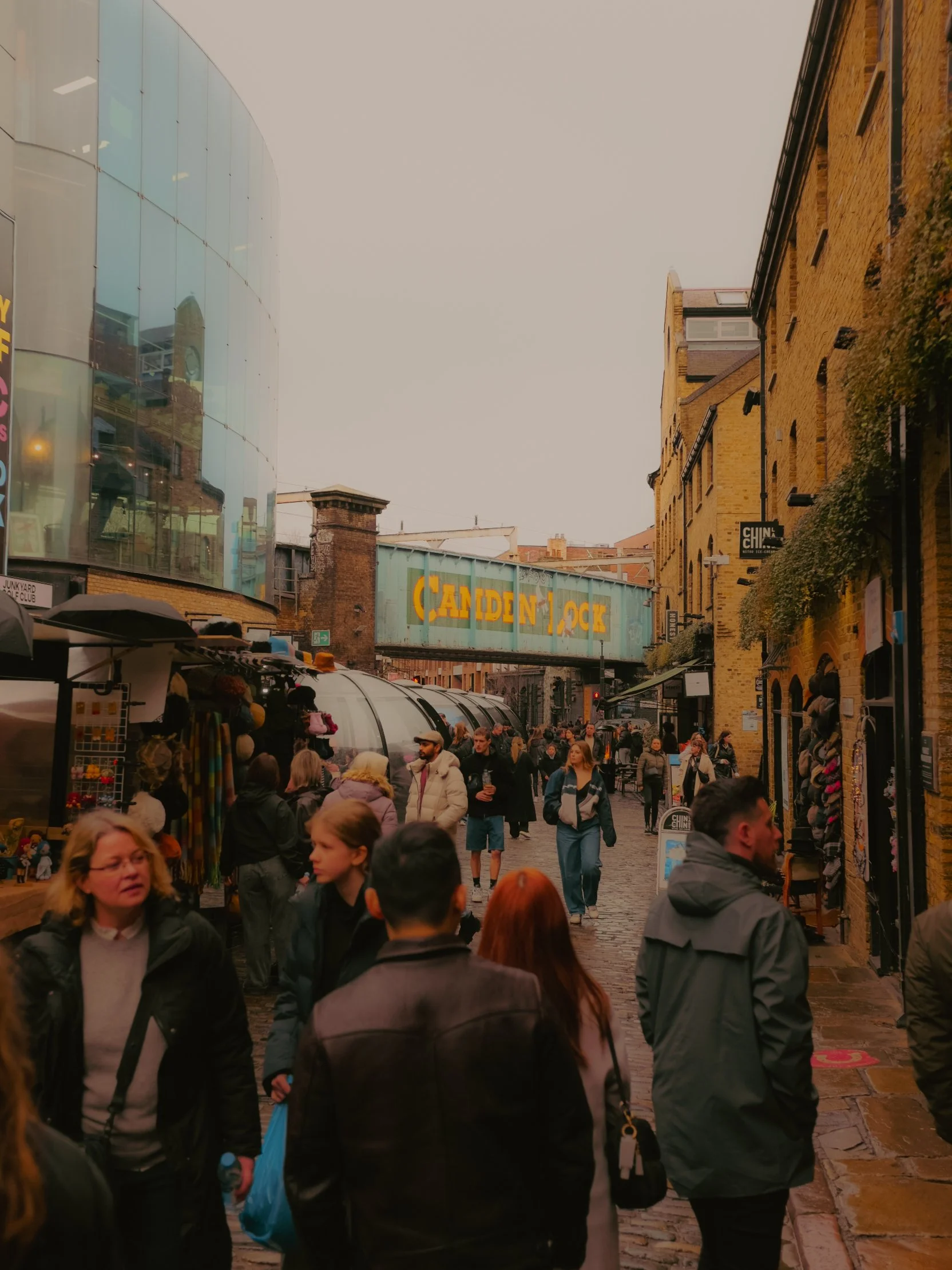 Rue animée avec des personnes de divers âges marchant, boutiques et un pont métallique portant une enseigne 'Camden Lock' dans un quartier urbain.