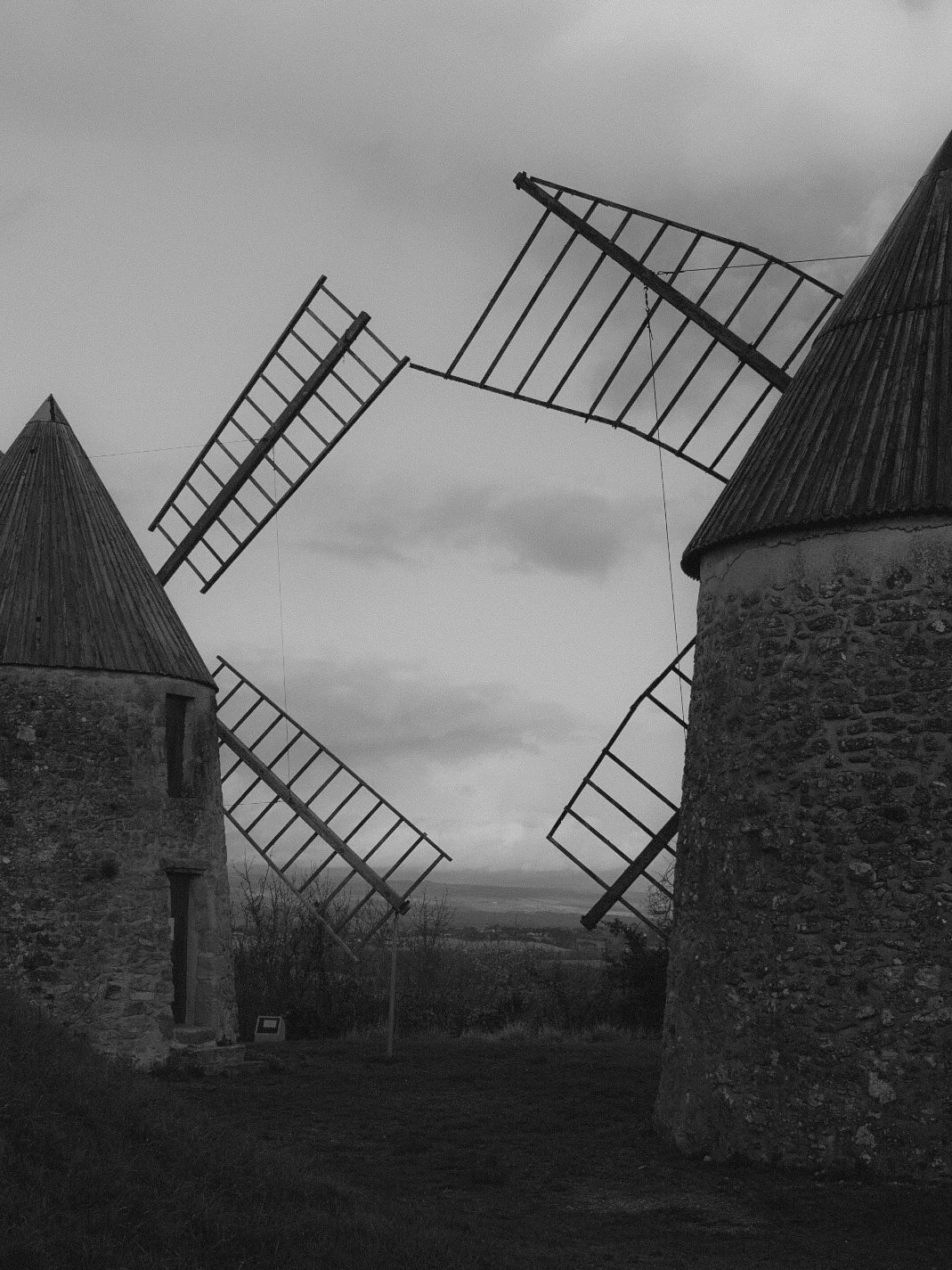 Deux moulins à vent en pierre, avec leurs ailes cassées ou déployées, dans un paysage rural sous un ciel nuageux.