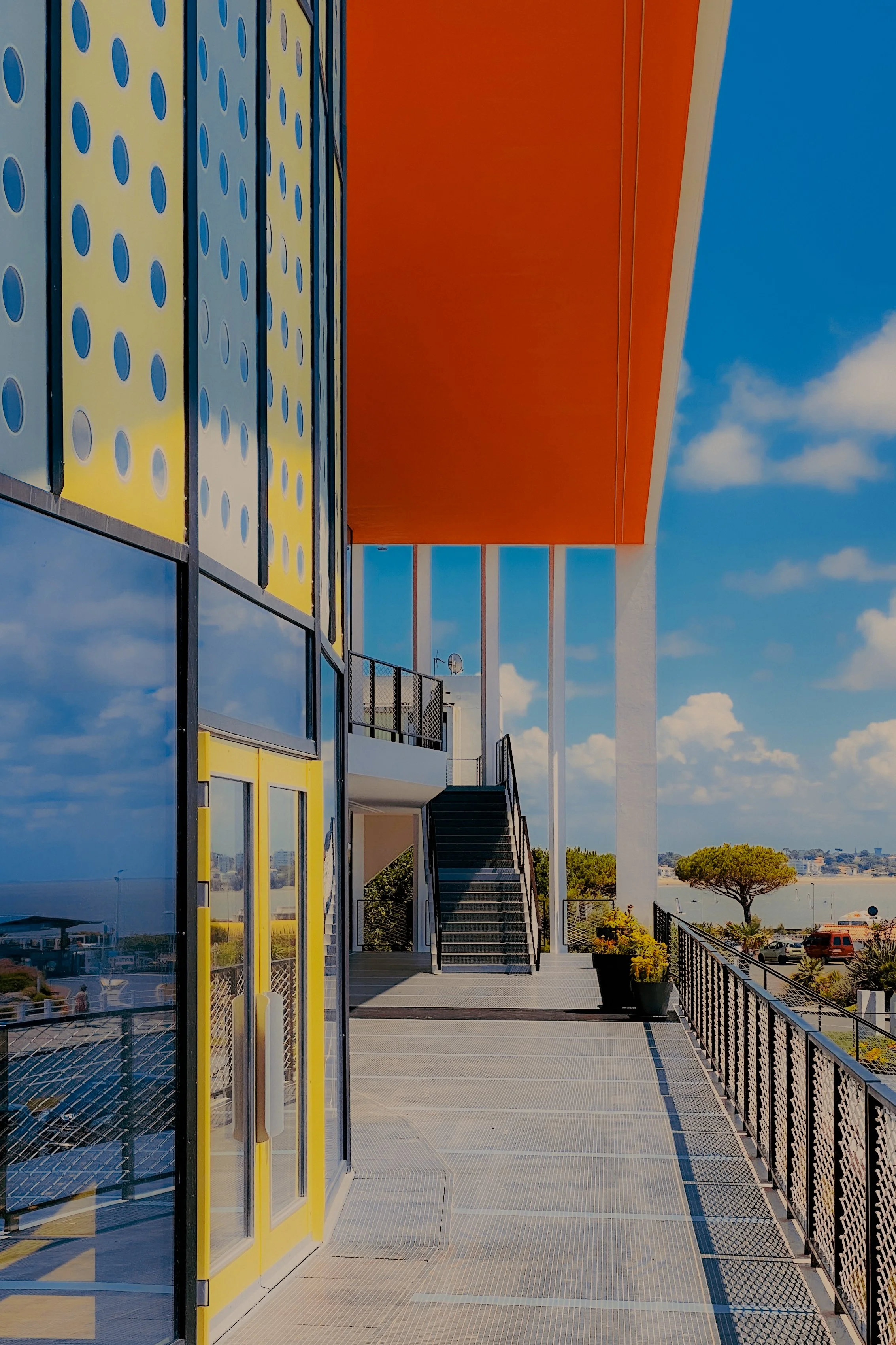Vue extérieure d'un bâtiment moderne avec façade en verre jaune et bleue, escalier menant à un niveau supérieur, balcon avec des plantes en pot, et ciel bleu avec quelques nuages.