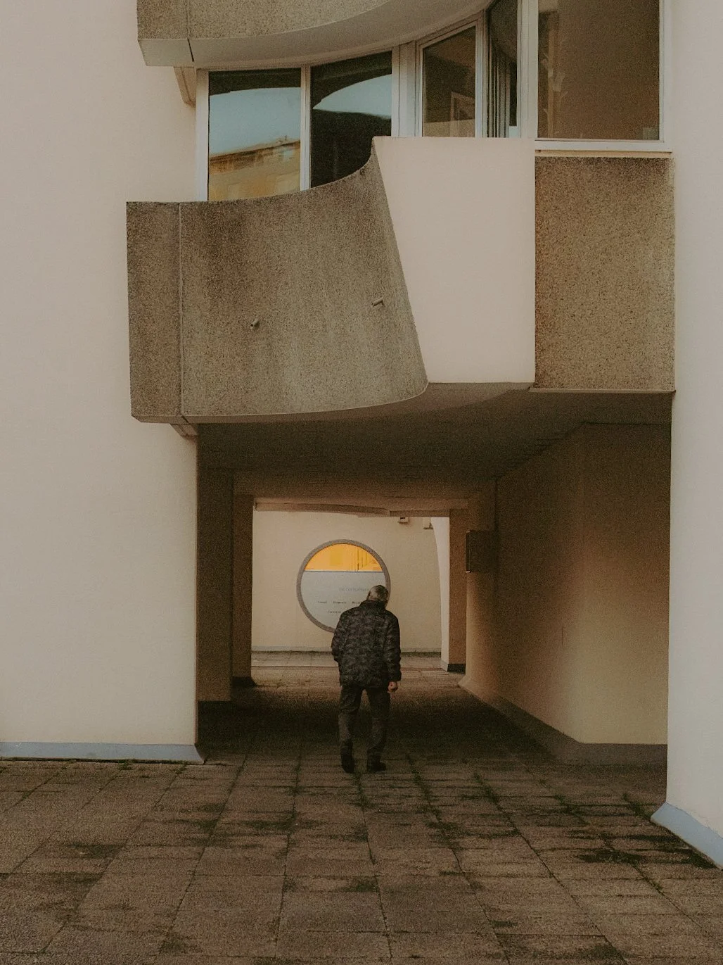 Un homme marche dans un passage couvert entre deux bâtiments modernes avec des balcons en béton en haut. À l'arrière-plan, une fenêtre ronde laisse passer la lumière.