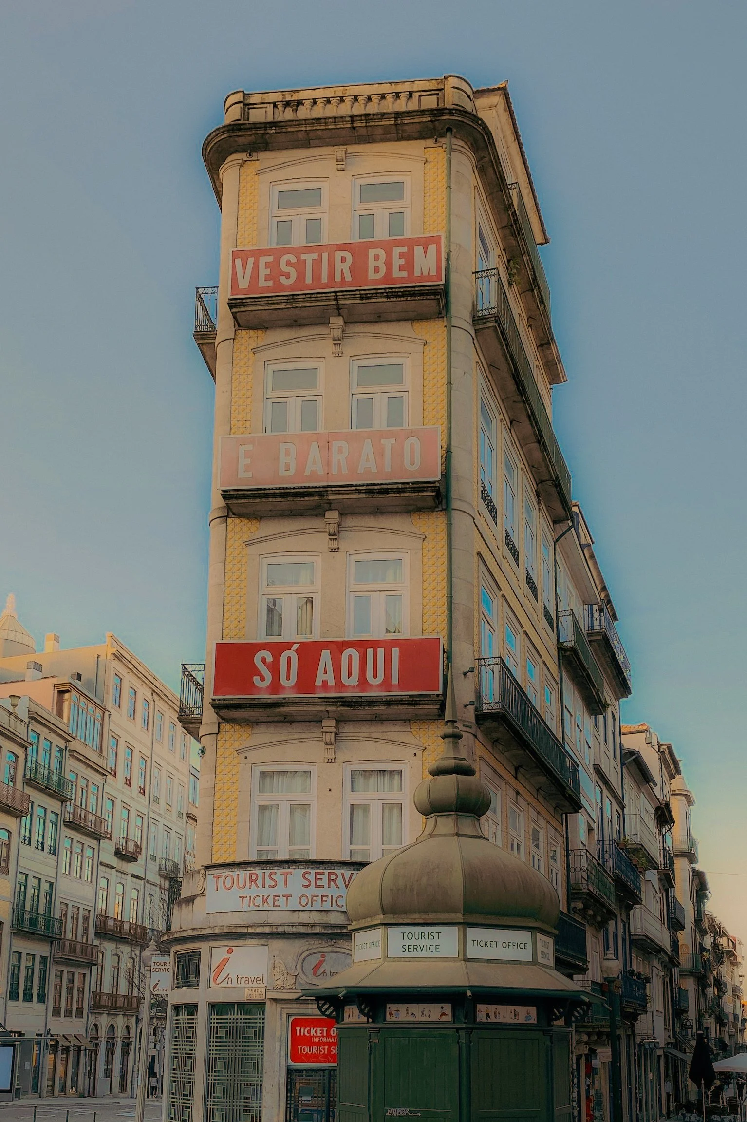 Un bâtiment ancien avec quatre panneaux rouges présentant un texte en portugais : « VESTIR BEM É BARATO » et « SÓ AQUI ». En bas, il y a un kiosque à tickets de service touristique. L'environnement urbain montre d'autres bâtiments anciens et une rue.