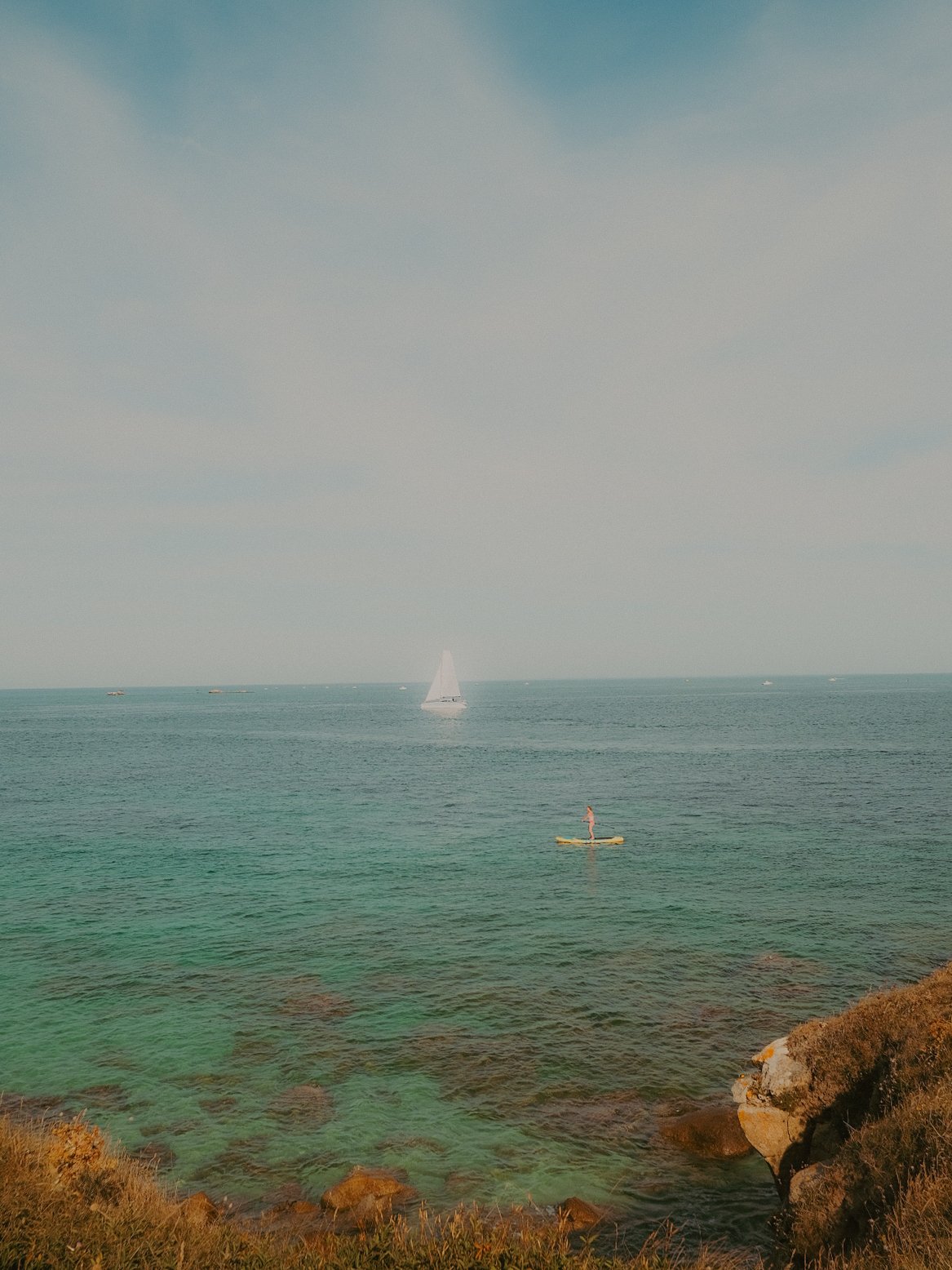 Une vue de la mer avec un voilier blanc, un paddle-boarder, des bateaux au loin, et des rochers avec de la végétation au premier plan.