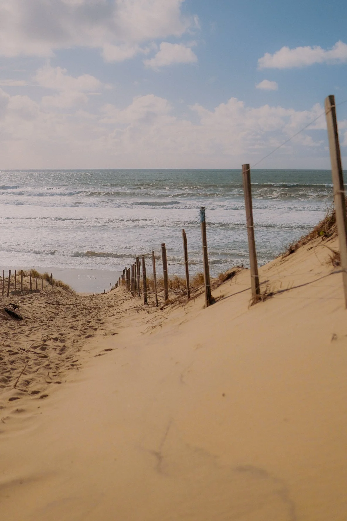 Sentier de sable menant à la plage avec des piquets de clôture en bois de chaque côté, dunes sableuses et océan avec vagues sous un ciel partiellement nuageux.