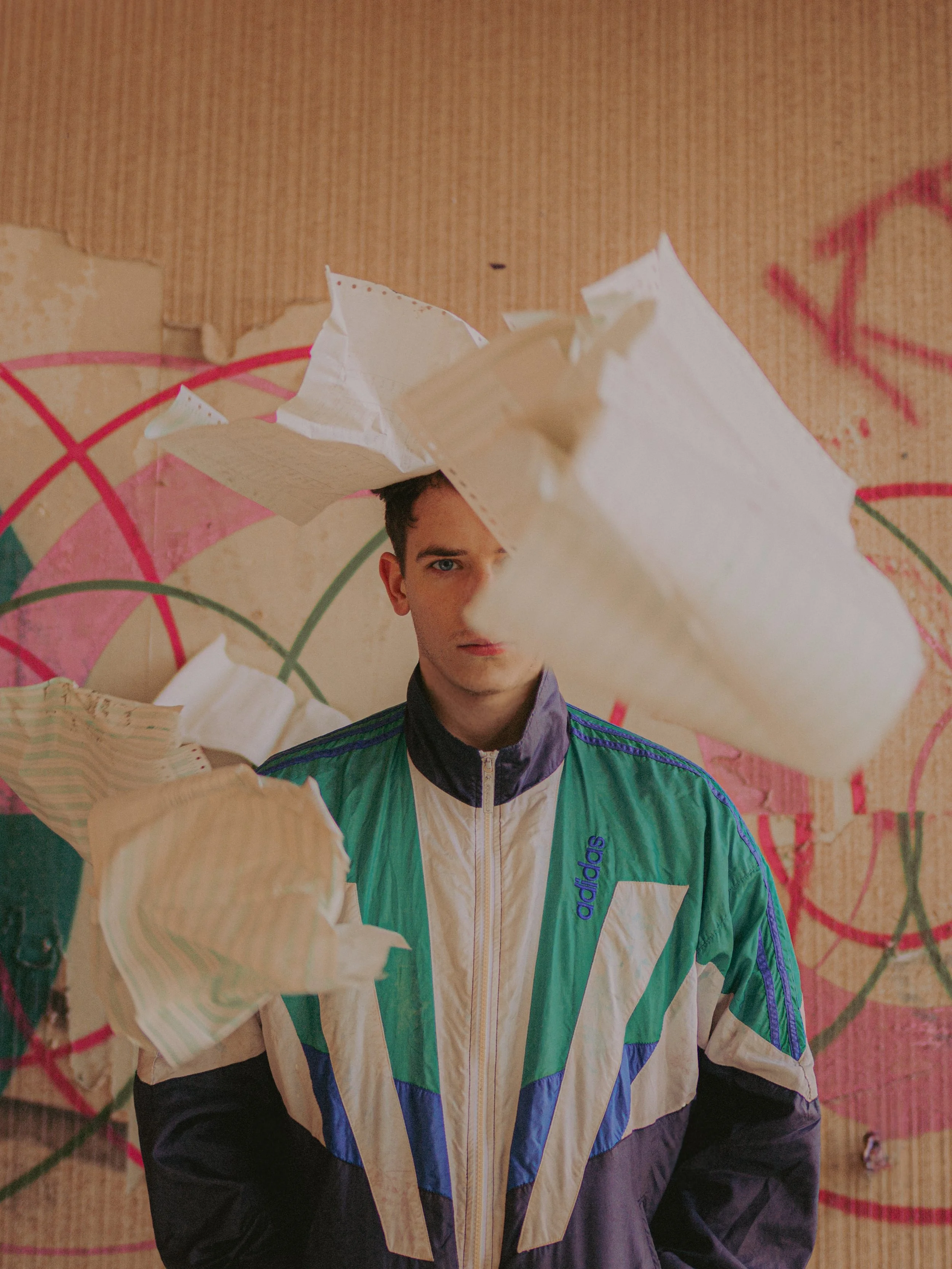 Un jeune homme portant un veston de sport coloré avec le logo Adidas, se tenant devant un mur avec des graffitis, entouré de feuilles de papier volant dans l'air.