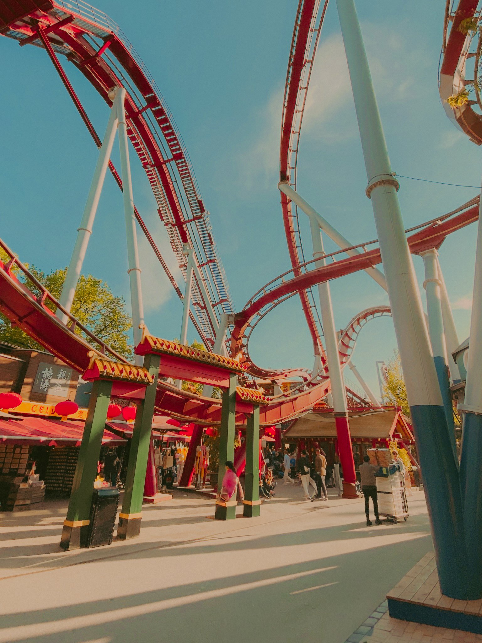 Vue d'un parc d'attractions avec une grande montagne russe rouge et blanche sous un ciel bleu, plusieurs visiteurs se promenant au sol, des structures décoratives de style asiatique et des lanternes rouges.
