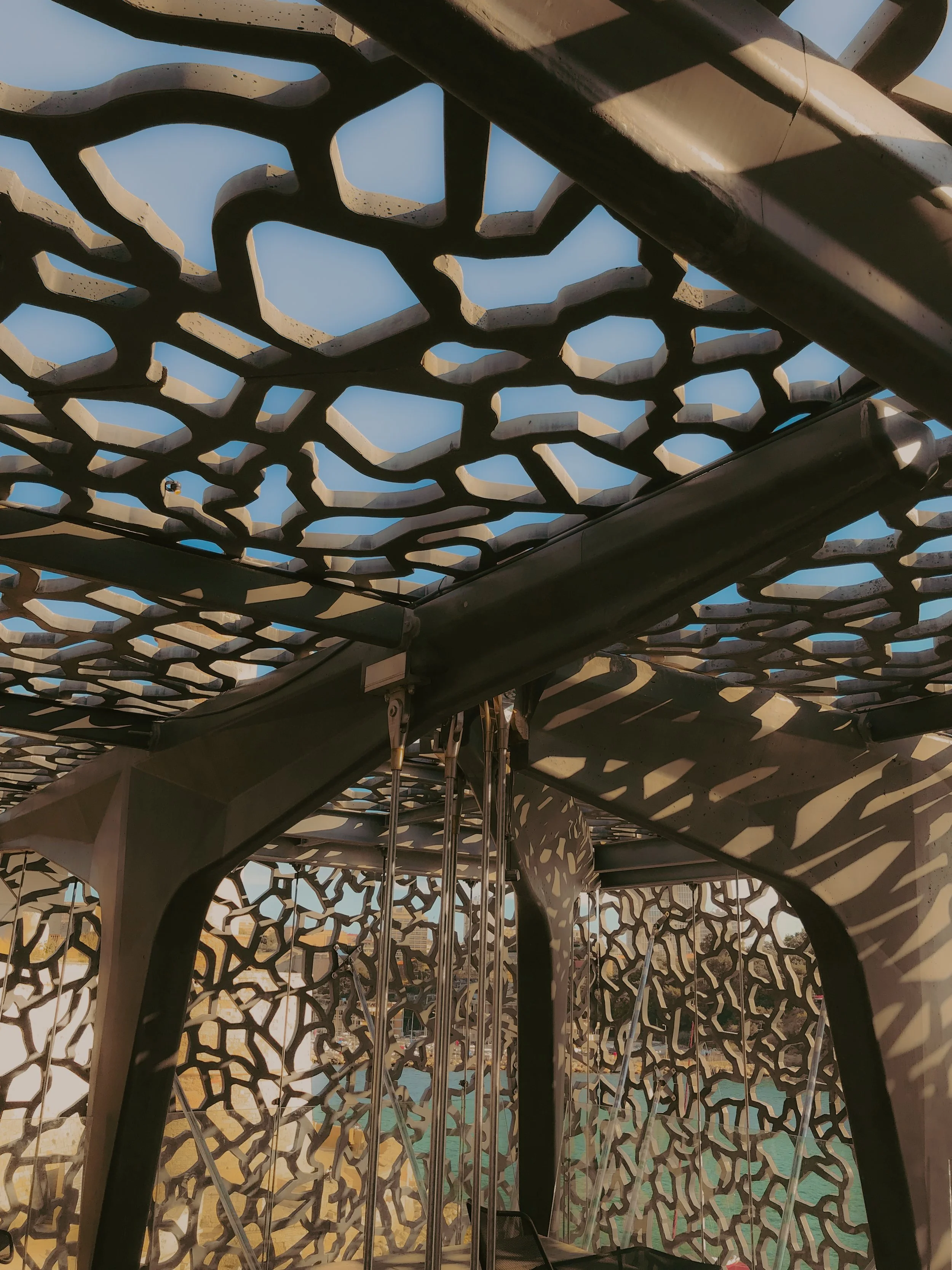 Structure moderne avec un motif complexe de découpes en forme de réseaux, laissant passer la lumière et créant des ombres. La structure semble être une partie d'un abri ou d'une parasol, avec des éléments en métal et un arrière-plan montrant une scèn